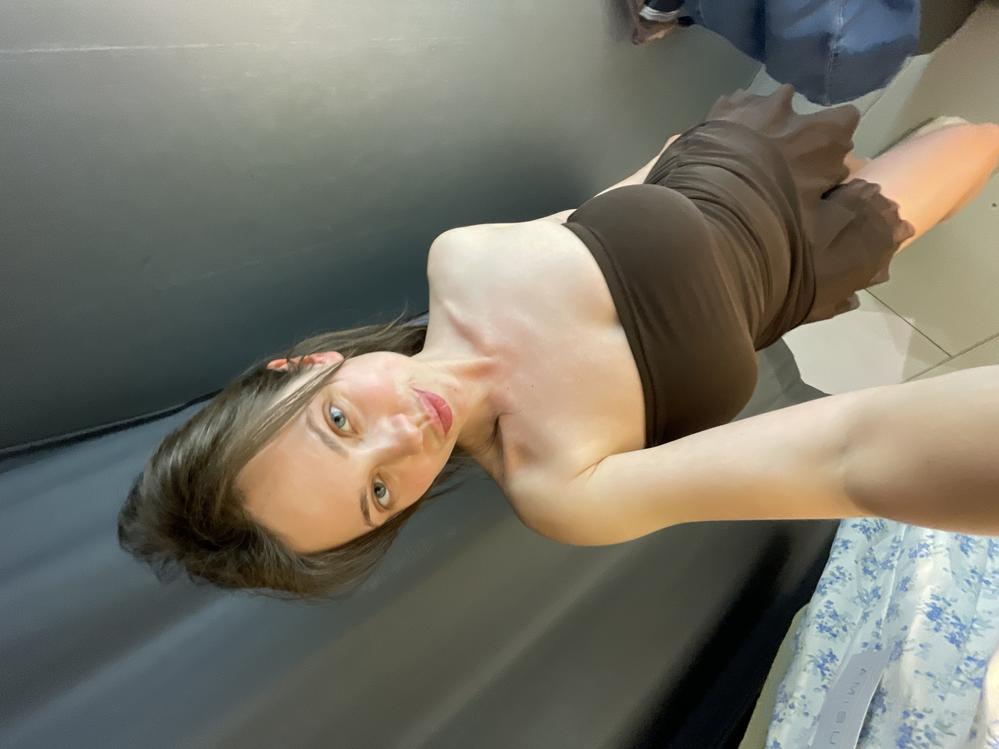 The image displays a full-body self-portrait of a young woman, captured indoors and rotated 90 degrees clockwise. The woman is positioned diagonally across the frame, showcasing a form-fitting, strapless, dark brown mini-dress with a ruffled skirt. She has long brown hair, striking blue eyes, and red lipstick, looking directly at the camera with a calm expression. Her left arm is extended, suggesting she is holding the camera for the selfie.
The background consists primarily of a plain, dark gray or black wall, indicating an enclosed indoor setting, possibly a fitting room. In the lower right quadrant of the image, a light-colored fabric with a blue floral pattern is visible, on which the brand name "AMISU" can be clearly read. Further towards the upper right, dark-colored items, possibly clothing or a bag, are partially visible.
The activity is a casual selfie, likely taken to display the dress. No specific time of day or weather can be determined from the indoor setting. The location context provided is Budapest, Hungary, though no specific visual cues within the image confirm this.