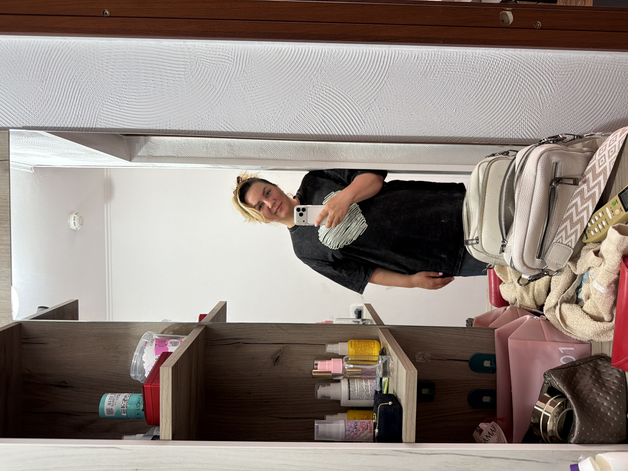 A woman with light blonde hair is holding a white smartphone, taking a selfie in a mirror. She is wearing a black t-shirt with a white design. The reflection shows a room setting with a light-colored wall and what appears to be a cabinet or shelf unit with various items: toiletries, cosmetics, bags, and other miscellaneous objects. The location is Leningradskaya, Russia. The lighting is bright, and the focus is on the reflection of the woman.