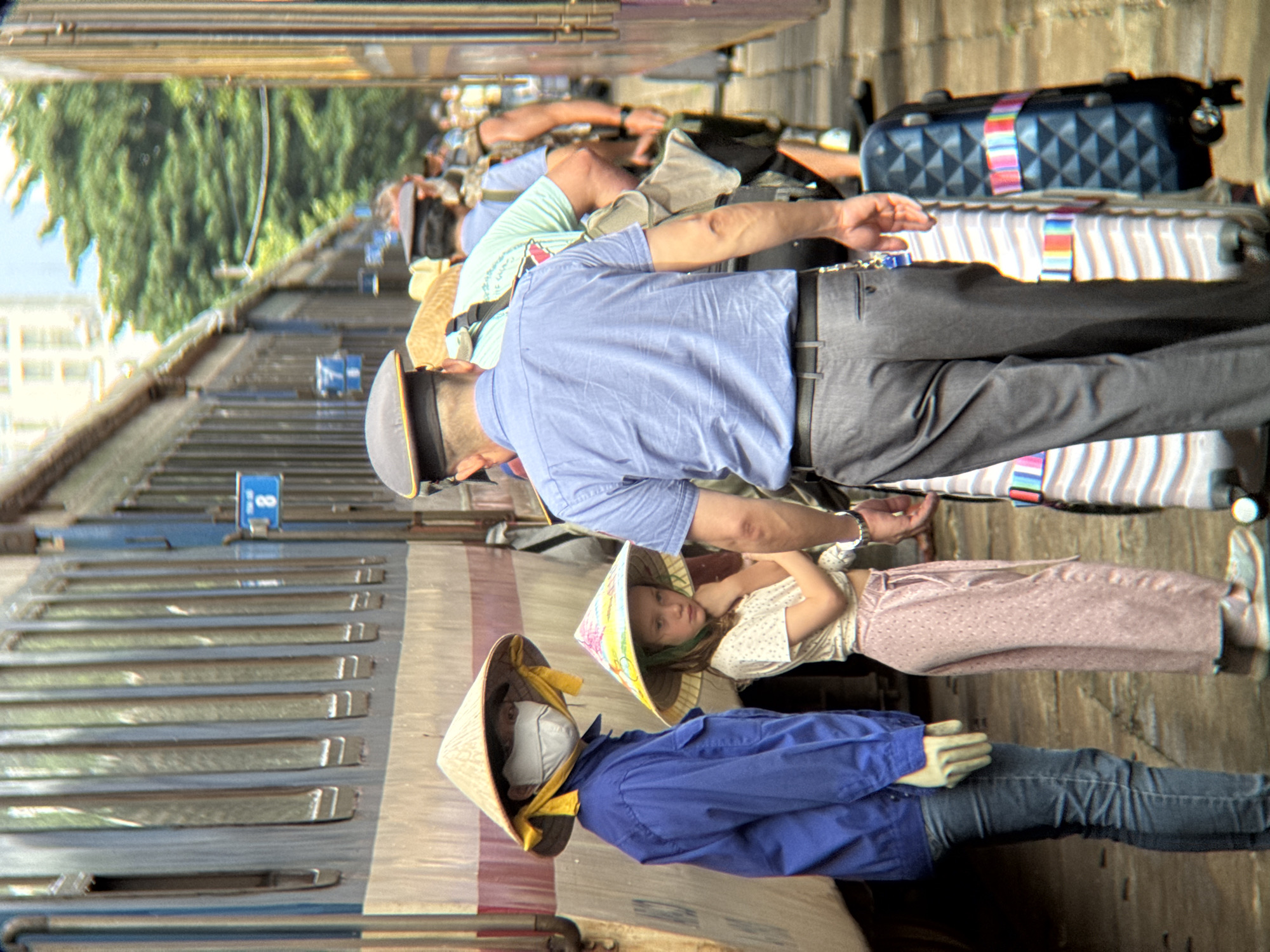 The image depicts a street scene in Da Nang, Vietnam, likely near a train station, with a train visible on the left. Several people are present, some wearing conical hats typical of Vietnam. In the foreground, a person in a blue shirt and jeans stands next to a young girl wearing a similar conical hat. Behind them, a man in a blue shirt and grey pants is seen from the back, with luggage beside him. Other individuals are visible further down the platform, some with luggage, suggesting a travel or departure scenario. The environment appears to be an outdoor, urban setting with a train and a paved walkway.