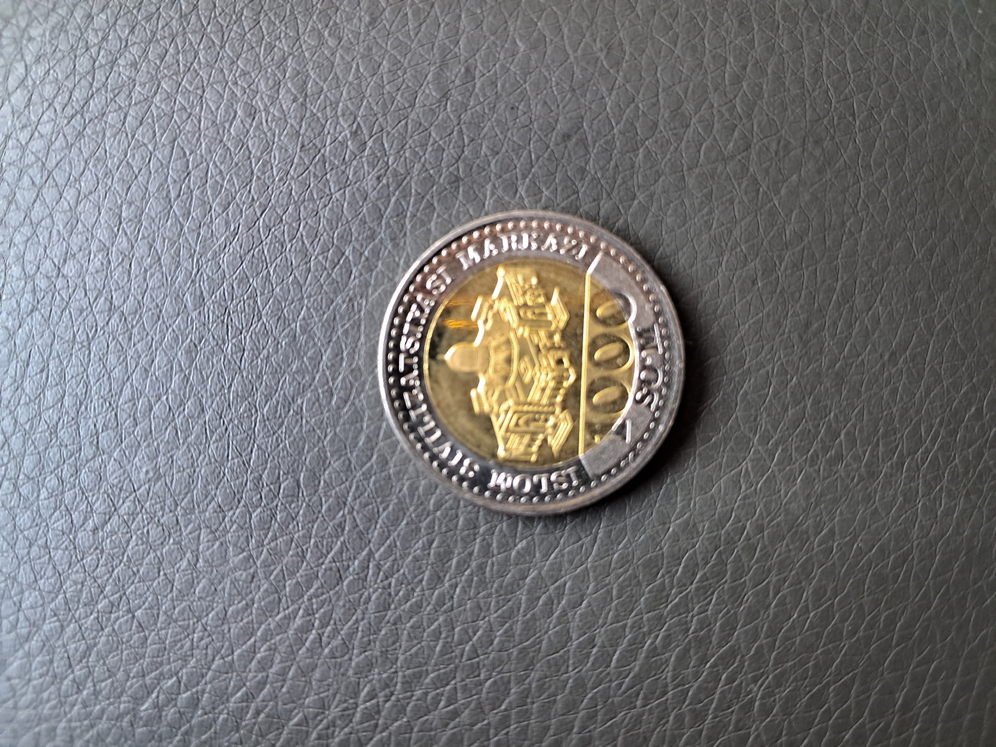 A coin rests on a dark, textured surface, likely leather. The coin has a gold-colored center featuring an embossed image of a building with domes and arches, possibly a mosque or historical structure. Around the central image, the coin has a silver-colored rim with raised dots and lettering. The lettering around the rim is partially obscured but includes what appears to be "UZBEKISTAN" and possibly other text. Below the central image, the denomination "2000" is clearly visible in gold, followed by "SUM" in silver. The overall impression is of a commemorative or currency coin. The location in the city of Bagish, Uzbekistan is not ascertainable from the image.