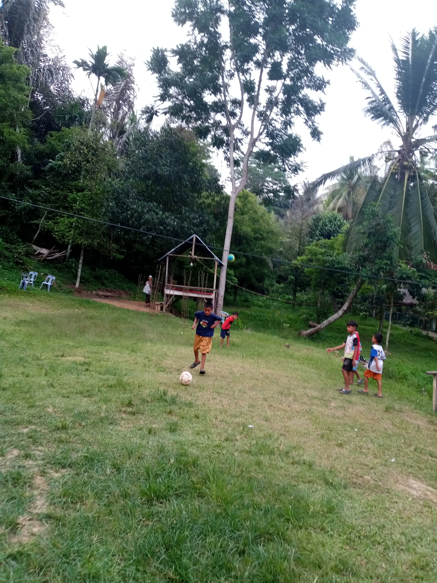 The image depicts an outdoor scene with several children playing soccer on a grassy field. In the background, there is a wooden structure resembling a gazebo or shelter, surrounded by lush vegetation and trees. The environment suggests a rural or semi-rural setting, possibly within the city of Lhokseumawe, Indonesia, given the presence of tropical flora. A few plastic chairs are visible to the left of the frame. The sky appears overcast.