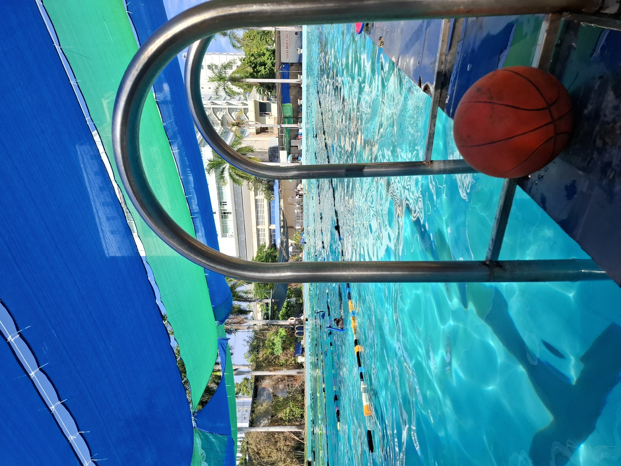 This image captures a vibrant outdoor swimming pool scene, likely located in Nha Trang, Vietnam, given the presence of palm trees and the warm, sunny atmosphere. The pool is a brilliant turquoise blue, with lane markers clearly visible, suggesting it's a public or communal facility. A set of metal ladder rails leads into the water, and a weathered orange basketball rests on the pool's edge, hinting at recreational activity.
Overhead, a blue and green tarp or shade cloth is stretched across, providing cover from the sun. In the background, several tall buildings, characteristic of a city environment, are visible, interspersed with lush green foliage and palm trees. The water surface is rippled, reflecting the sunlight and the overhead coverings, creating a dynamic interplay of light and shadow.
A few individuals can be seen in the pool, some appearing to be swimming or snorkeling, adding a sense of life and activity to the scene. The overall impression is one of a relaxed, sun-drenched day at a well-maintained swimming facility in a tropical urban setting.