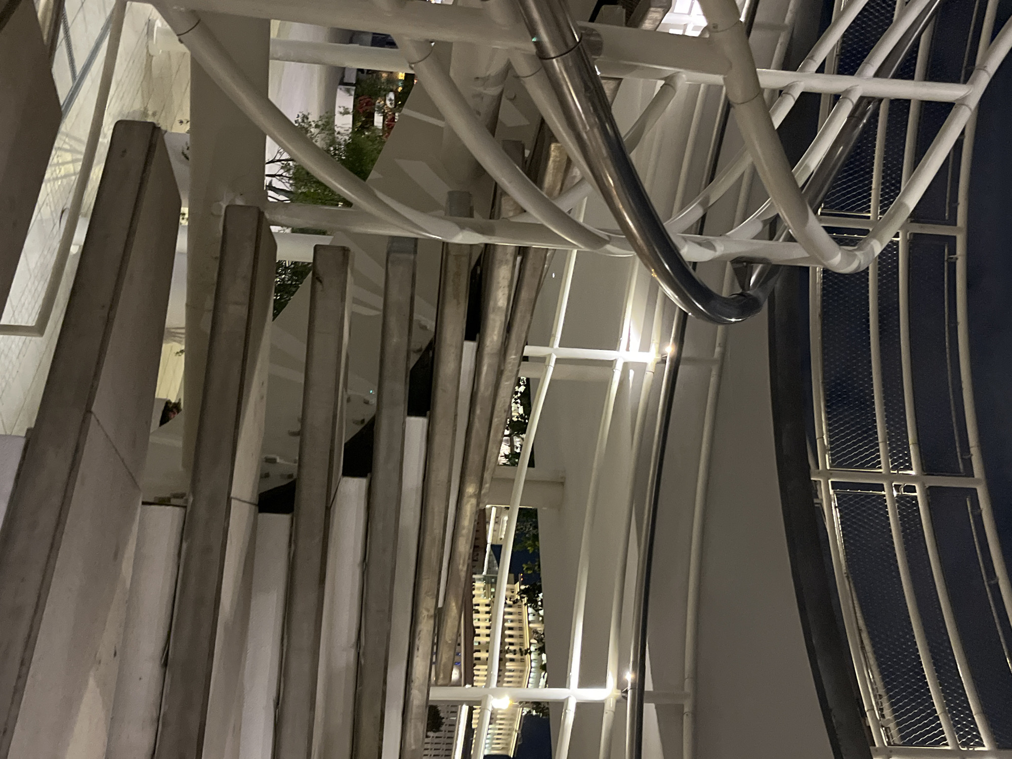 This media file presents an abstract, architectural shot of a complex structure at night. The image, oriented sideways, primarily features a dense network of white and grey architectural elements.
In the foreground, a series of light grey, rectangular beams or slats run vertically across the left side of the frame, creating a rhythmic, textured pattern. Behind and interwoven with these are numerous thick, curved white pipes or supports that bend and intertwine, forming an intricate, almost sculptural design. A prominent, reflective silver-colored pipe also curves through the white structure.
Further into the background, sections of a white metal mesh fence are visible against a dark expanse, which appears to be the night sky. Peeking through the gaps in the structure are glimpses of distant, artificially lit buildings and dark green foliage, indicating an urban setting.
The time of day is clearly night, with various light sources illuminating the structure, highlighting its contours and casting subtle shadows. No people or specific activities are visible. The overall impression is one of modern, complex design, possibly part of a bridge, building facade, or a large-scale art installation. There is no visible text in the image. The location, according to context, is Moscow, Russia.