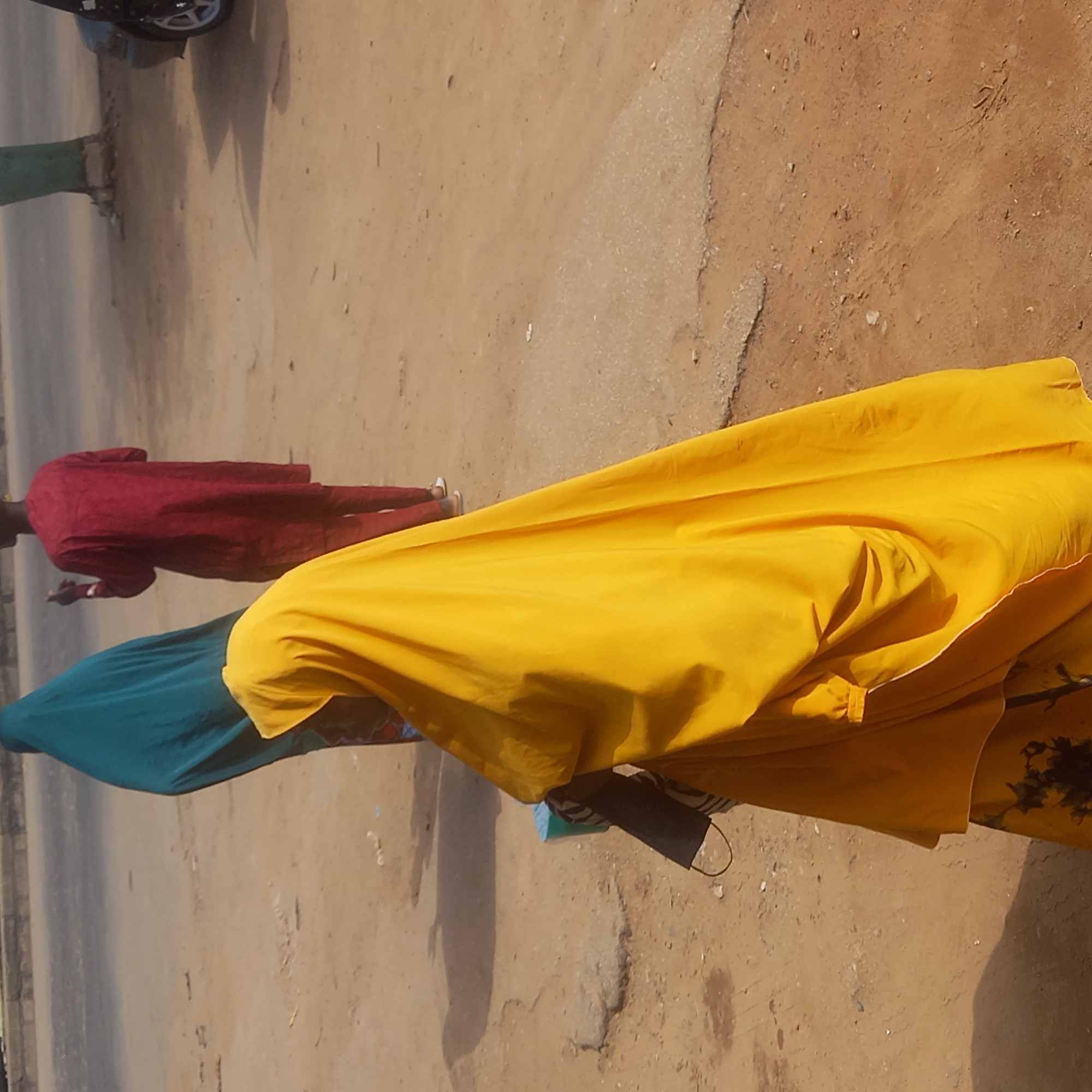 The image shows three people on a dirt road in Sardauna, Nigeria. One person is wearing a red outfit and white shoes. A person is wearing a blue outfit, and the closest person is wearing a yellow outfit. There is a dark face mask hanging off of the yellow outfit. There is also a parked motorcycle in the background.