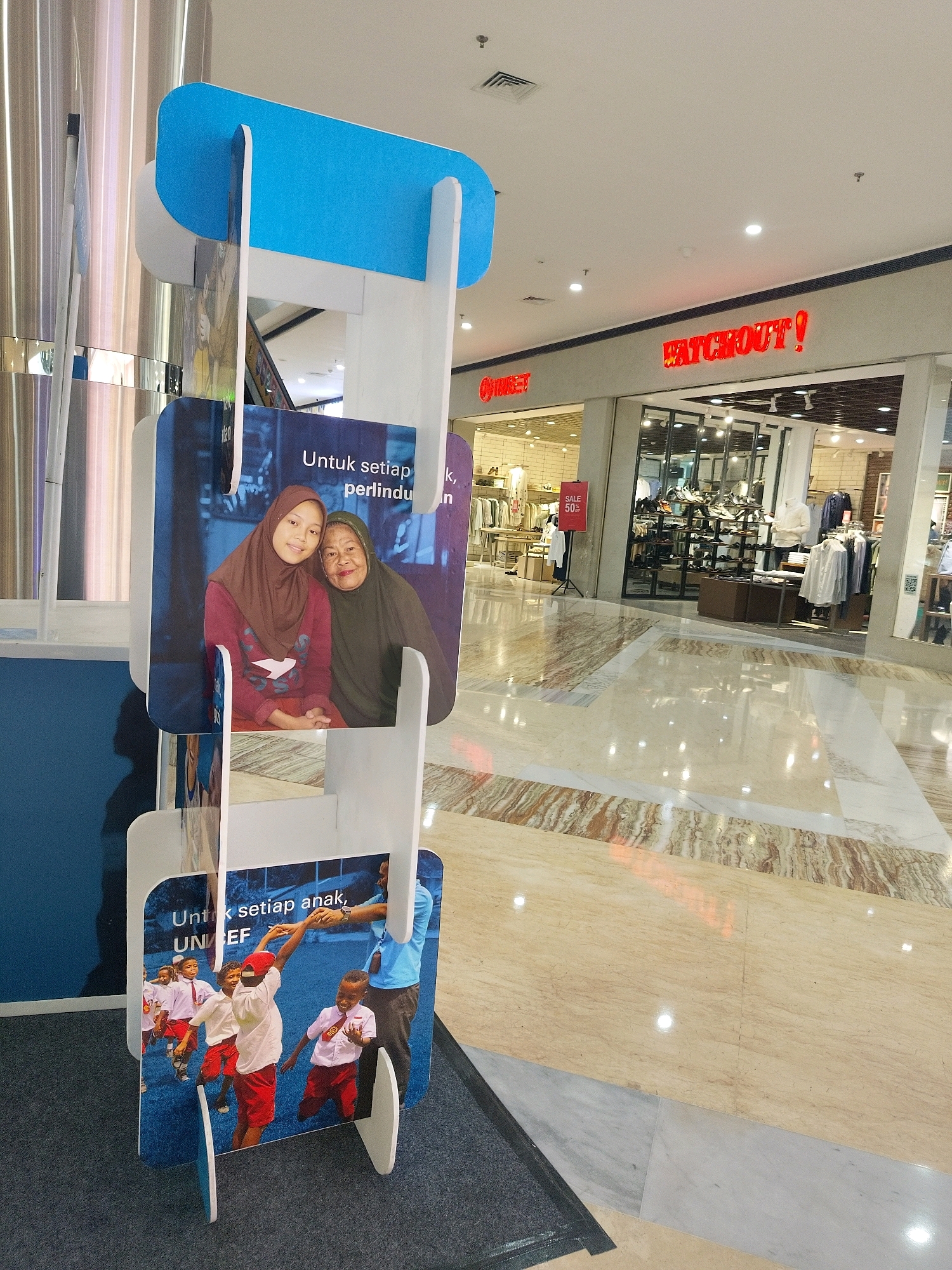 The image shows a display stand with three panels, likely in a shopping mall in Mojokerto, Indonesia. The top panel is blue and white. The middle panel features a photograph of two women, one young and one elderly, wearing hijabs and smiling. Text in Indonesian, "Untuk setiap anak, perlindungan," which translates to "For every child, protection," is visible on this panel, along with the UNICEF logo. The bottom panel displays a photograph of a group of young boys in school uniforms playing and running, with an adult in the background. The floor is polished marble with a patterned design. In the background, a store with the red neon sign "WATCHOUT!" is visible, along with clothing displays and mannequins. A "SALE 50%" sign is also present in the background. The lighting suggests an indoor environment, likely a mall.