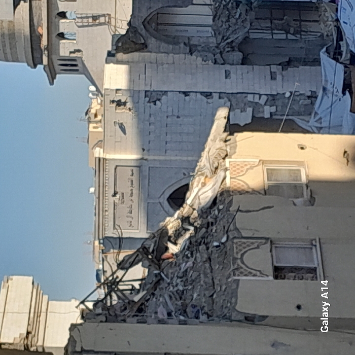 The image displays a scene of extensive structural damage in Khan Yunis, Palestinian Territory, under a clear blue sky. In the foreground, the upper section of a building, characterized by cream-colored walls and decorative arched frames above two rectangular windows, has collapsed into a significant pile of rubble, exposing twisted metal rebar. Behind this debris, the central structure is identified as the Mosque of Sheikh Hammad Abu Khater Al Sheikh by Arabic script ("مسجد الشيخ حماد أبو خاطر الشيخ") visible above an arched entrance. This mosque and adjacent structures are heavily damaged, with large sections of walls crumbled and missing. To the right, a light-colored minaret, cylindrical with an octagonal upper section and small arched windows, stands partially visible, also showing signs of distress. White tarpaulins are discernible amidst debris in the lower right portion of the frame. No individuals or active movements are visible.