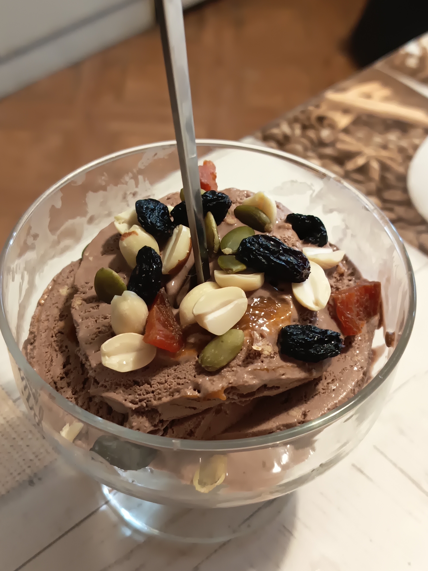 A close-up, eye-level shot shows a serving of chocolate ice cream in a clear glass bowl, topped with various ingredients. A metal spoon is inserted into the ice cream. The toppings include peanuts, raisins, pumpkin seeds, and pieces of dried fruit, possibly apricots. The ice cream itself appears to be a rich brown color and has a slightly melted texture. The glass bowl is clear and has a stem. The background is blurred but suggests an indoor setting with wooden surfaces and hints of a patterned placemat or tablecloth. The image does not provide any specific indicators of its location in the city of Izmail, Ukraine.