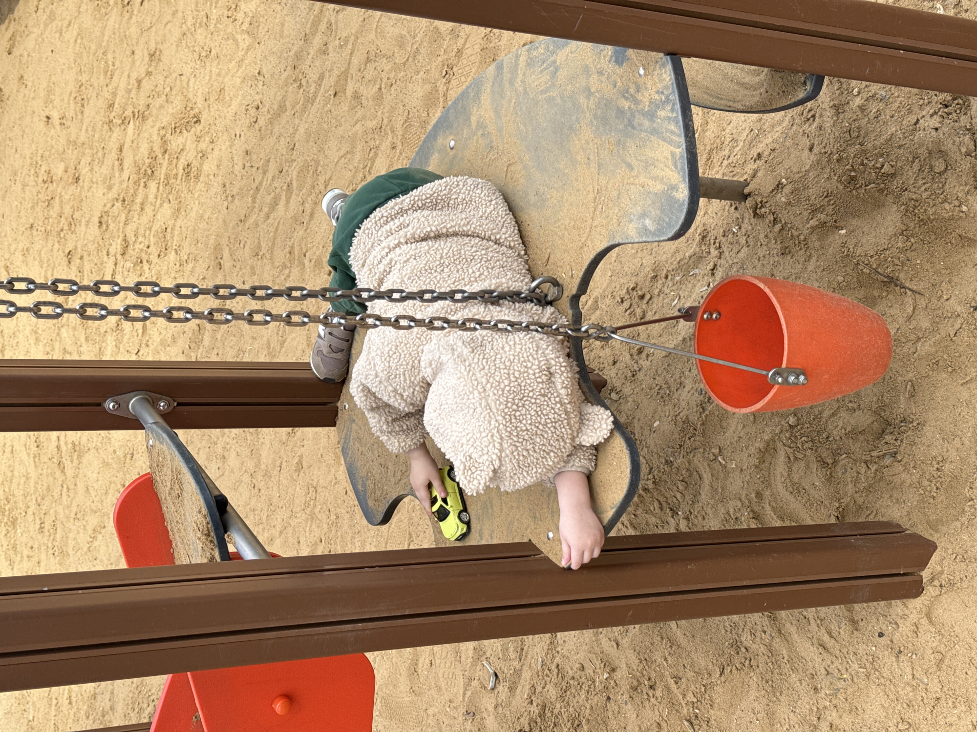A child wearing a cream-colored, sherpa-like jacket and green pants is playing on playground equipment in what appears to be a sandbox. The child is situated on a piece of equipment with a gray, irregularly shaped seat. The child is holding a small, yellow toy car with their hands. An orange bucket hangs nearby, attached by a metal chain and rigging. The playground equipment is bordered by brown wooden beams and red metal pieces. The location is likely a playground with a sandbox area. Moscow, Russia is not evident in the image.