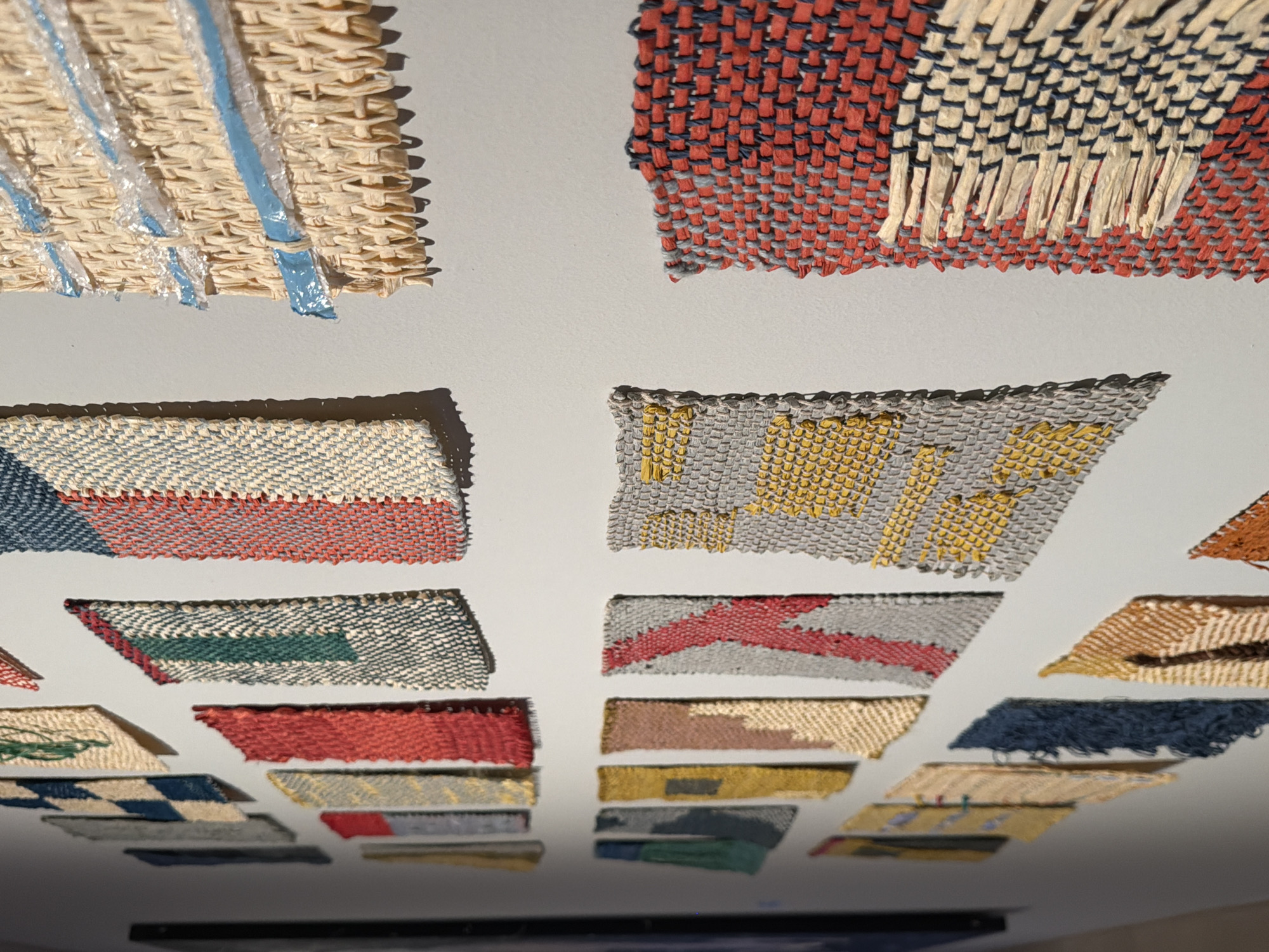 The image shows a collection of woven textile samples displayed on a white wall. The samples are of various sizes, colors, and patterns, including geometric designs, stripes, and color blocks. Some samples have a fringe or frayed edges. The lighting is even, and the samples cast slight shadows on the wall. There is no clear indication of location. The image does not provide enough information to determine if it is located in Tel-Aviv, Israel.