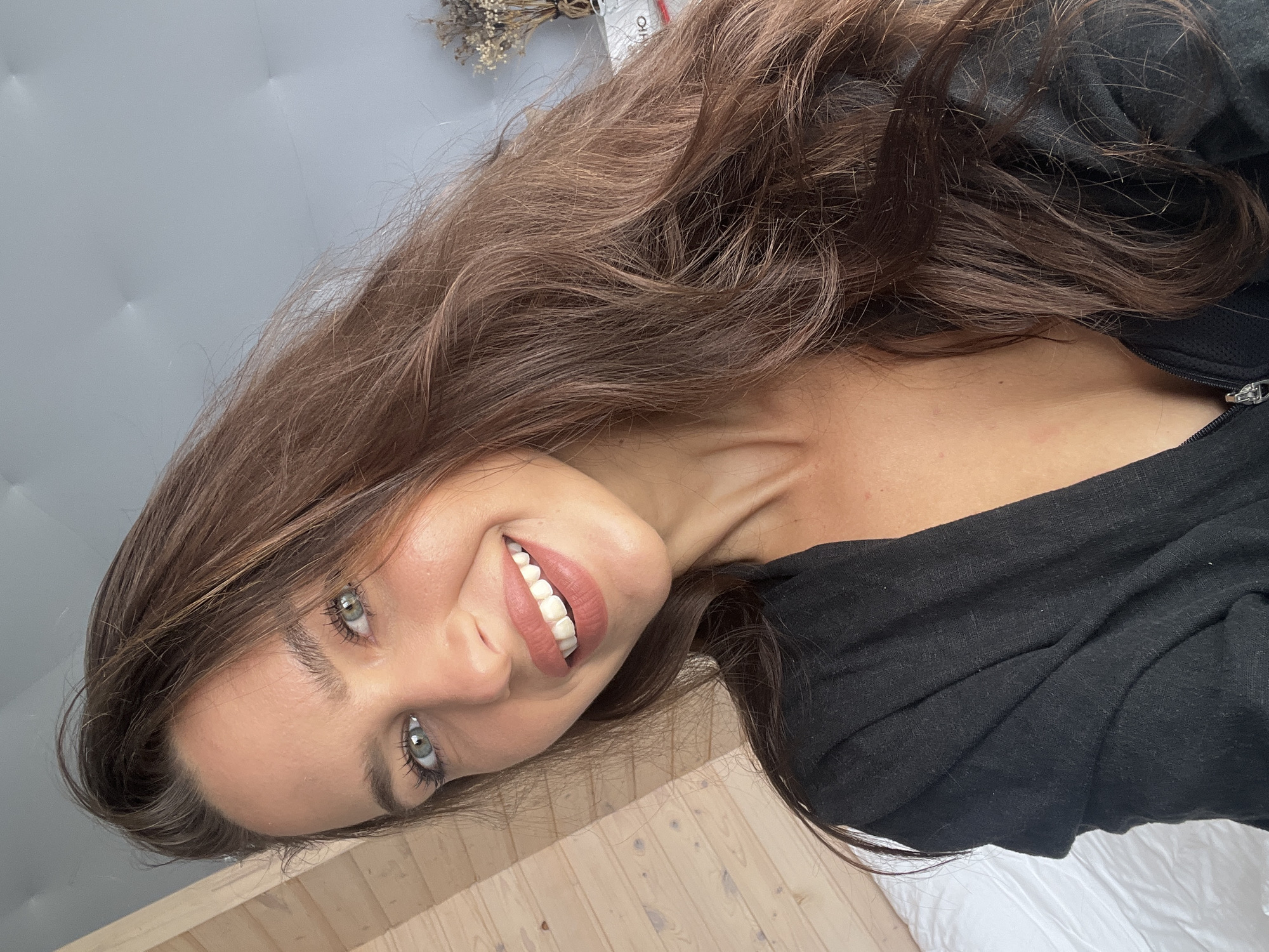The image is a selfie of a woman with long brown hair, light skin, and green eyes. She is smiling, revealing white teeth. She is wearing a black garment, possibly a shirt or jacket. The background includes a light gray padded headboard and light-colored wooden floorboards. A dried floral arrangement is visible in the upper left corner. The image does not contain any information or elements suggesting a location in the city of Zarubino, Russia. The description of a location in Zarubino, Russia is not determinable by visual elements.