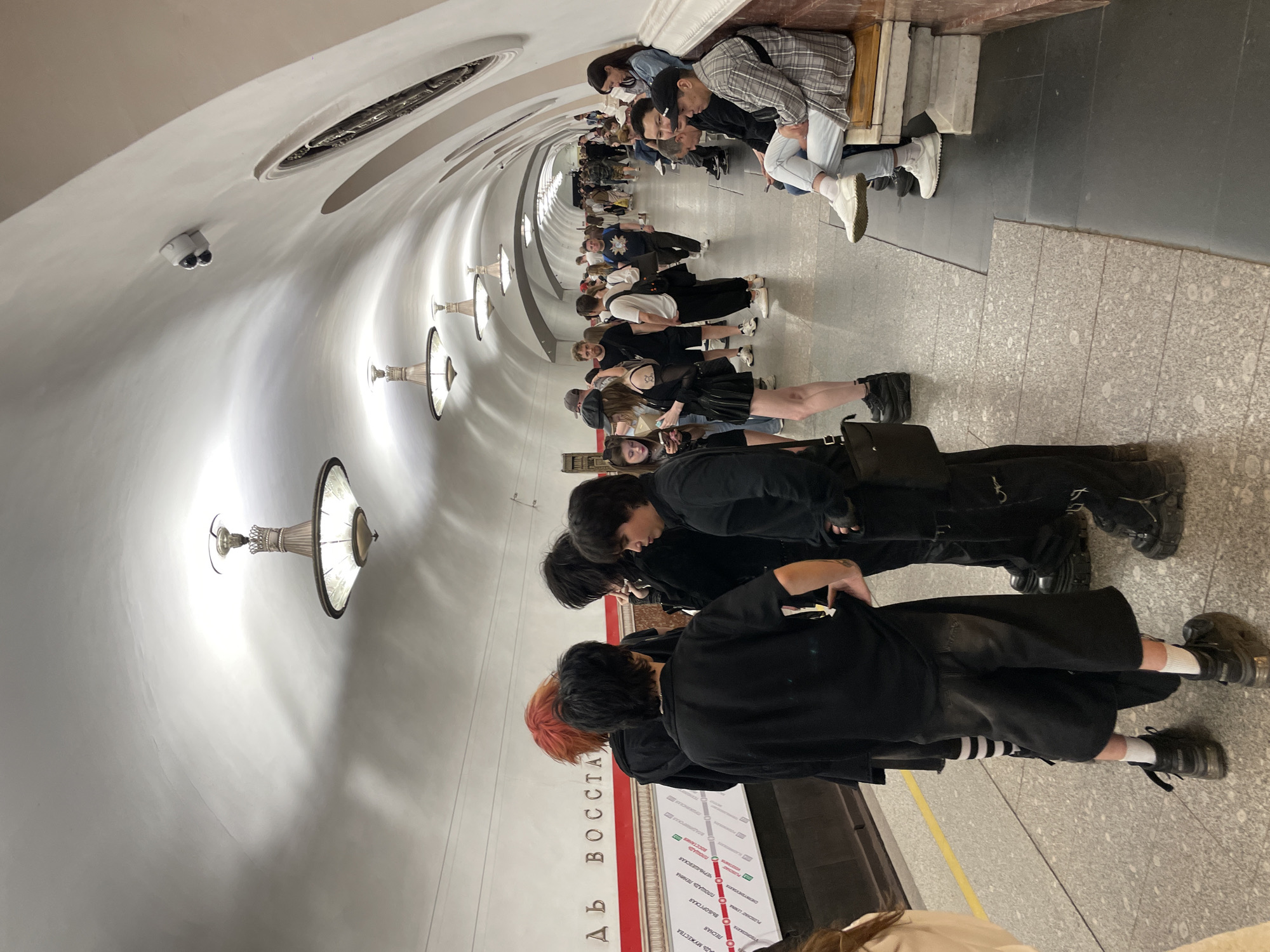 The image depicts the platform of Ploshchad Vosstaniya metro station in Saint Petersburg, Russia. The station features a high, vaulted white ceiling illuminated by multiple ornate chandeliers with glass shades. A white security camera with two lenses is mounted on the ceiling in the upper right quadrant. The platform floor is primarily light gray textured tiles, with a darker gray section bordering the right wall. A thin yellow line is visible on the platform near the tracks on the left.
On the left wall, a white sign with a red horizontal stripe displays Cyrillic text, partially visible as "АДЬ ВОССТА" (likely "Ploshchad Vosstaniya") and a subway line map indicating stations such as "ПЛОЩАДЬ МУЖЕСТВА," "ПЛОЩАДЬ ЛЕНИНА," and "ЧЕРНЫШЕВСКАЯ."
In the foreground, a group of five individuals with alternative fashion styles is standing. From left to right: a person in black clothing with white and black striped socks partially visible; a person in a black t-shirt, black wide-leg pants/shorts, and black chunky sneakers; a person with bright orange-red hair wearing a black hoodie; a person with long dark hair wearing black clothing; and another person in black clothing with very chunky black boots, a small black bag over their shoulder, and a visible tattoo on their right arm. This group appears to be interacting, with one individual looking down towards a phone held by another.
In the middle ground and background, numerous other passengers are present. Some are walking along the platform, extending into the far distance of the tunnel, while others are standing. On the right side, several people are seated on a light-colored wooden bench positioned against a dark reddish-brown marble wall. This wall curves upward, transitioning into the white vaulted ceiling, and features decorative circular elements. One man seated on the bench wears a plaid shirt and light blue jeans with white sneakers. A woman in the middle ground on the left is observed looking at her mobile phone.