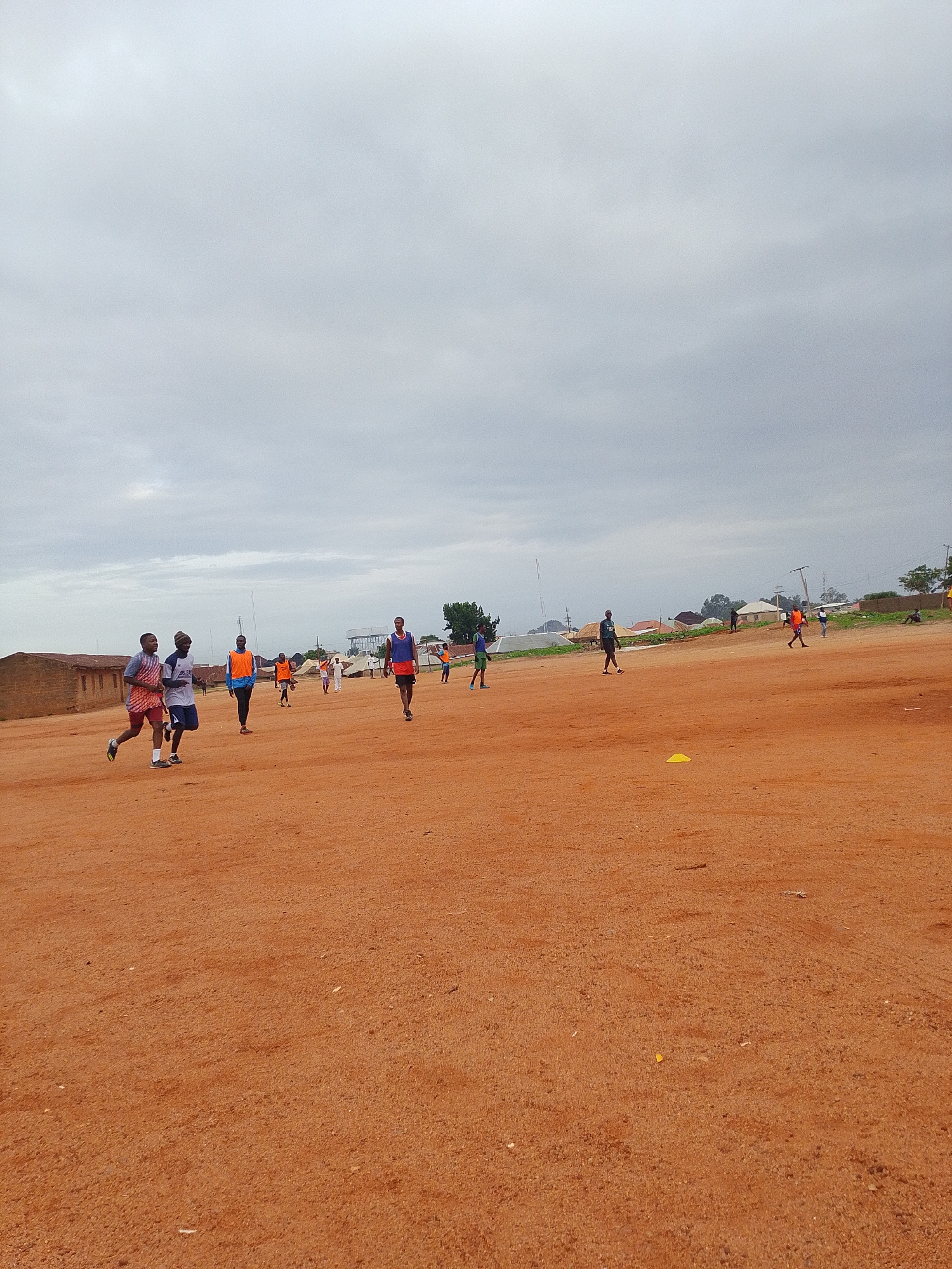 Location: Unknown, Unknown.
The scene depicts a group of individuals on a reddish-brown, dirt field. Several individuals are running or walking. Most are wearing athletic clothing, some with orange vests. The background shows a cloudy sky, buildings, and vegetation. A small yellow cone is visible on the field.