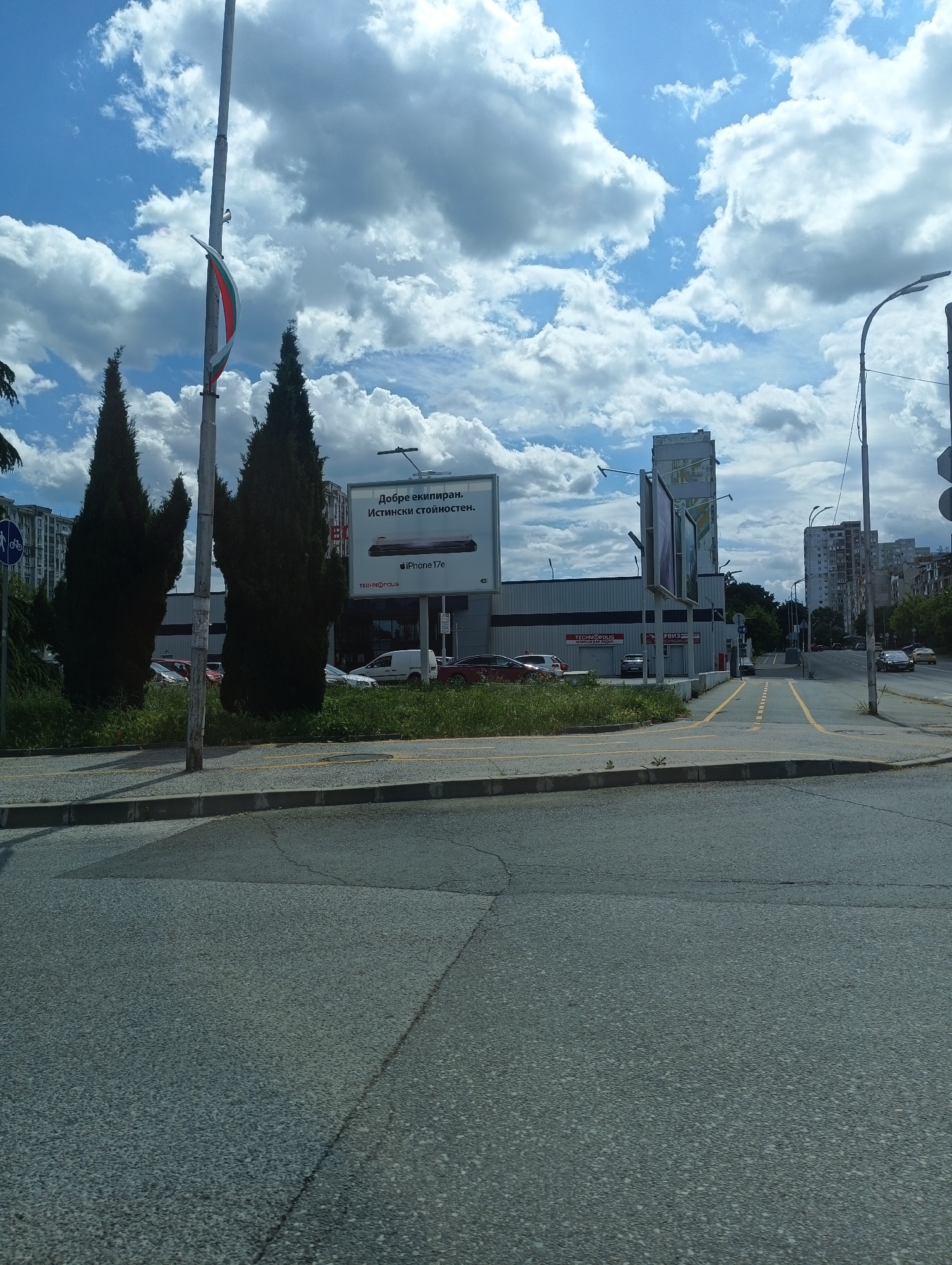 The image shows an urban street scene in Burgas, Bulgaria. The foreground is dominated by an asphalt road with minor cracks and pavement markings. On the left side of the road, there are tall, dark green conifer trees and a flagpole flying the Bulgarian flag. Behind the trees, there's a large billboard advertising an iPhone 17e. Behind that, a long, gray building with the name "TECHNOPOLIS" visible on the facade. Cars are parked in front of the building. In the background, there are residential apartment buildings and a partly cloudy sky. There are also some streetlights on the right side of the road.