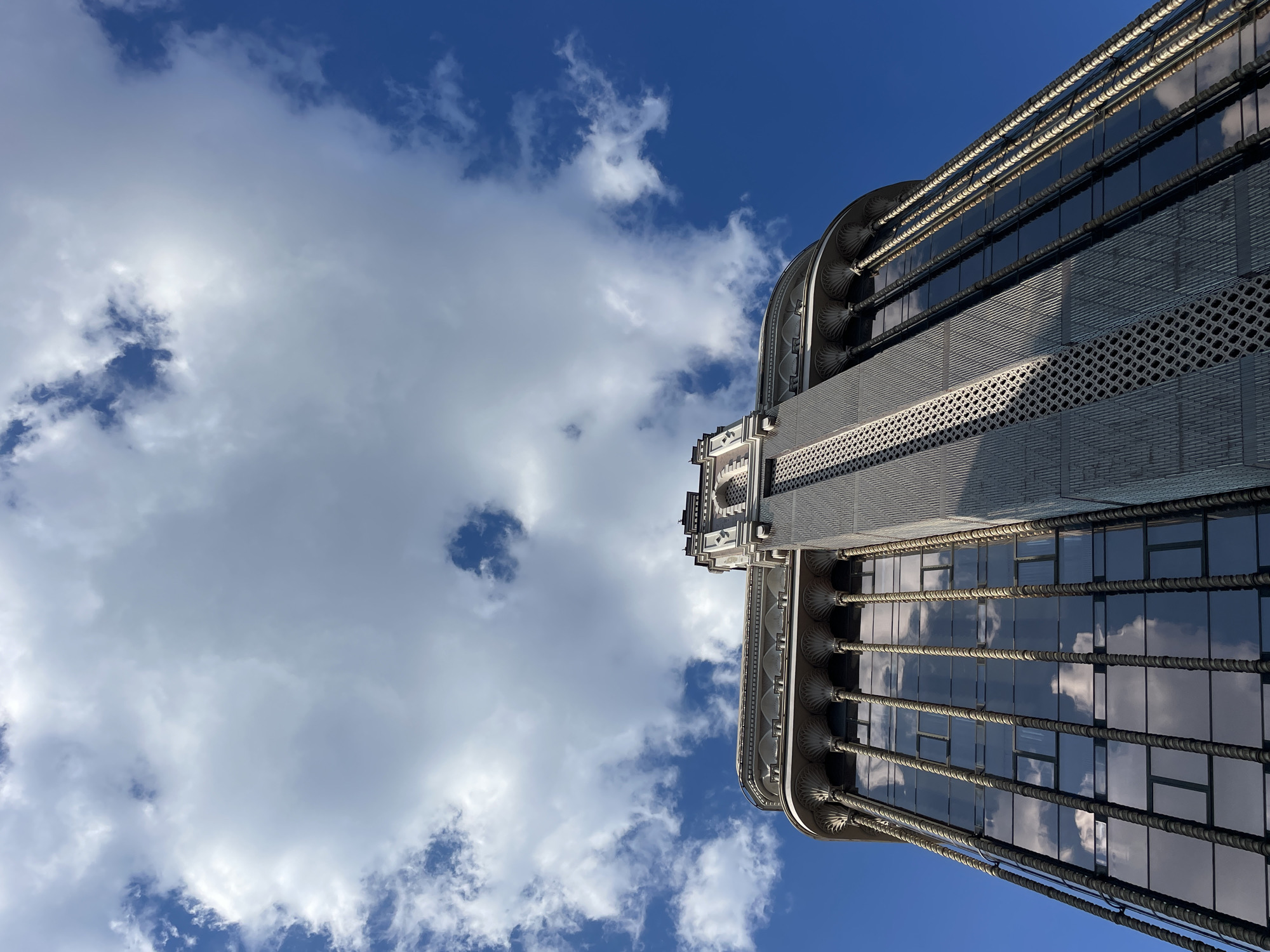 A low-angle shot shows a building rising into a blue sky with scattered cumulus clouds. The building has a modern design with repeating window units, vertical support structures, and decorative elements along its edge. The building reflects the sky and clouds, suggesting a glass facade. The location is unknown.