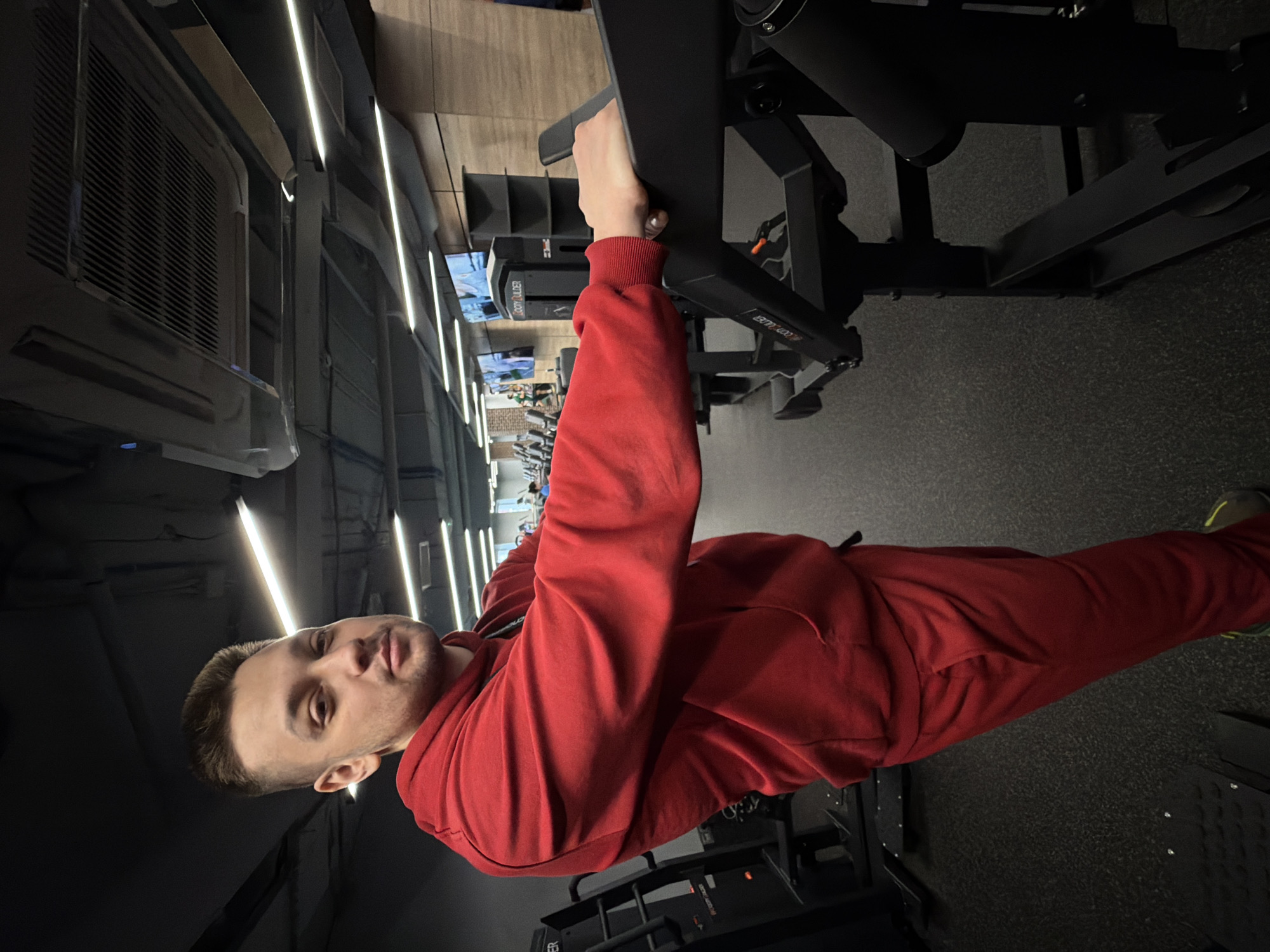 A man is shown in a gym setting, dressed in a red tracksuit. He's holding onto a piece of gym equipment. The gym has a modern appearance with dark flooring and equipment, and bright, linear lighting fixtures on the ceiling. A row of screens or monitors is visible in the background. The location is not identifiable as Saint Petersburg, Russia.