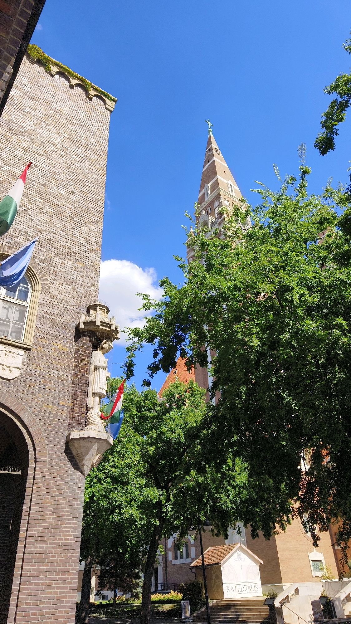 This is a photograph of the exterior of the Votive Church of Szeged, Hungary, on a clear day. The church is a prominent landmark in the city, known for its distinctive tower and facade. The image captures the church's intricate architectural details, including its brickwork, spires, and stained-glass windows. In the foreground, lush green trees partially obscure the view of the church, creating a sense of depth and framing the structure. Hungarian flags are visible, indicating a sense of national pride and celebration. The sky is a vibrant blue, with a few scattered white clouds, suggesting a pleasant summer day. The overall atmosphere of the photograph is one of peace and serenity, inviting viewers to appreciate the beauty of this historic religious site.