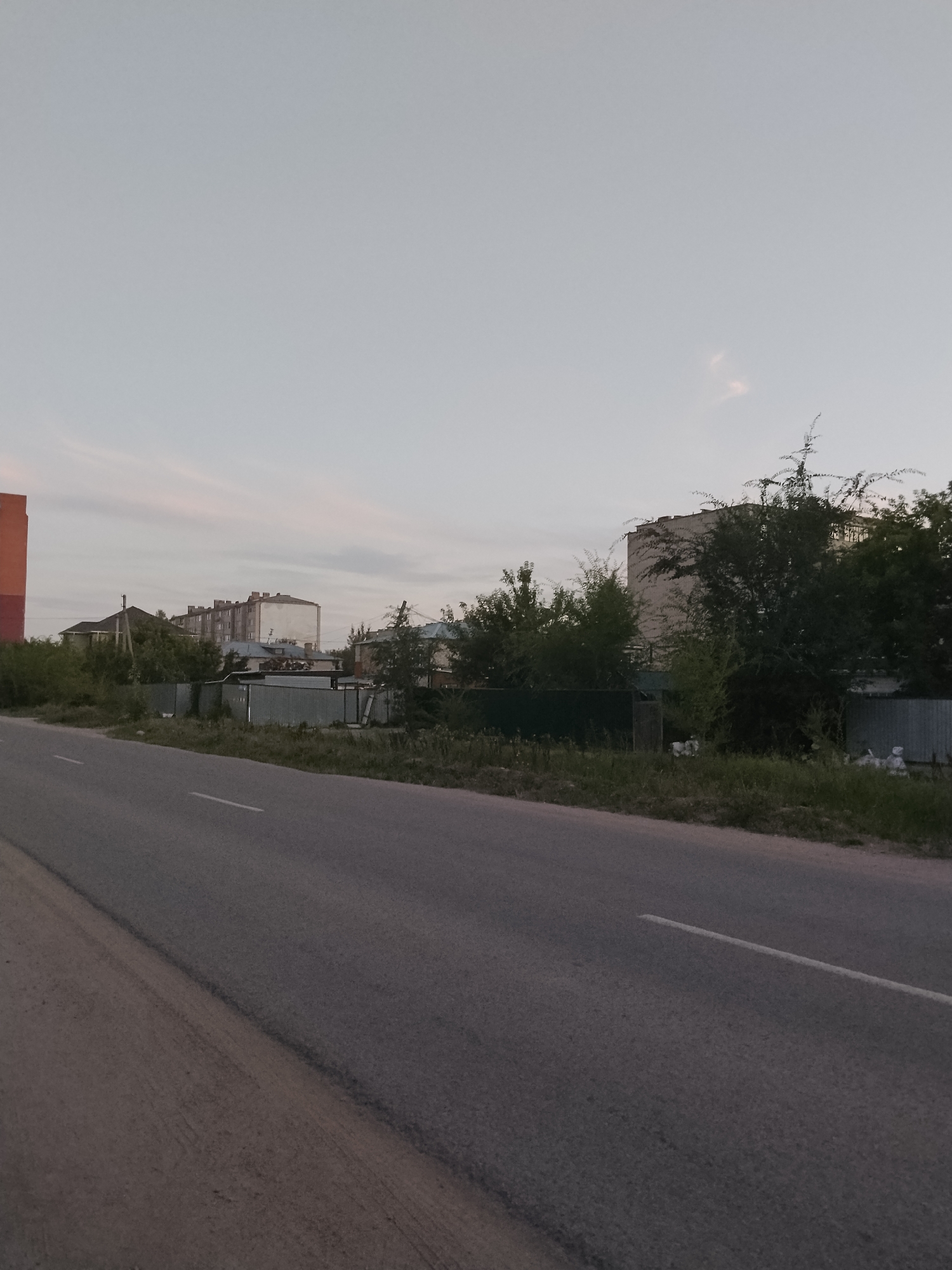 The image depicts a road in the city of Kokshetau, Kazakhstan, with residential buildings and vegetation along the roadside. The road is asphalt with a white dashed line marking the lane. On the right side of the road, there is a grassy verge with some trees and bushes. Beyond the vegetation, there are several fences, some made of corrugated metal and others of darker material. Behind the fences, there are residential buildings of varying heights and architectural styles. Some of these buildings appear to be multi-story apartment blocks with a neutral facade, while others are smaller, possibly individual houses. The sky is overcast with a pale, washed-out light, suggesting it is either early morning or late evening. There are no people visible in the image. The scene is one of a typical urban or suburban environment.
