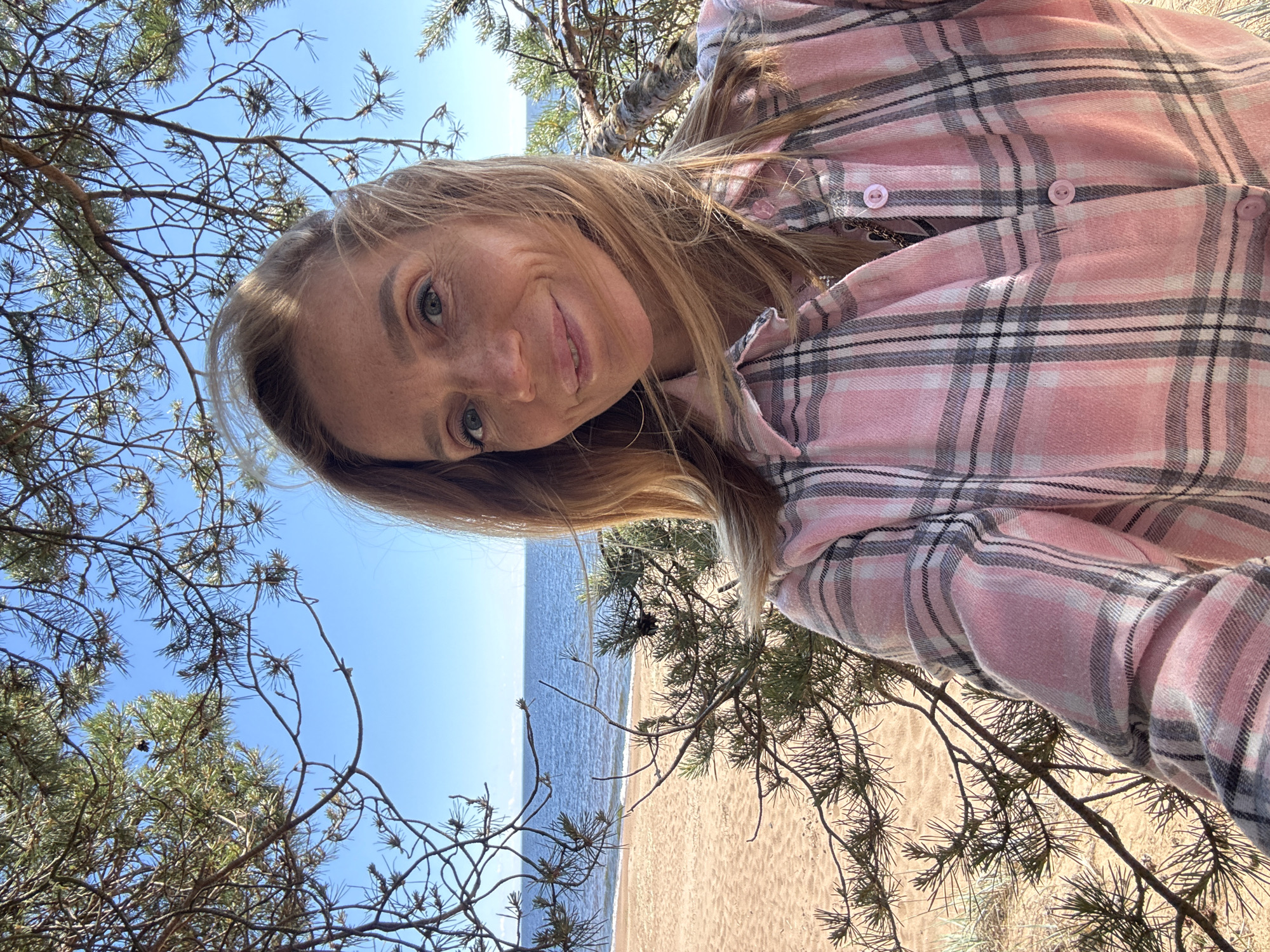 A woman is captured in a selfie taken outdoors. She is wearing a pink and grey plaid shirt. The background reveals a beach with sand and the ocean under a clear blue sky. Pine trees frame the image, with their branches and needles creating a natural border. The woman is smiling and looking at the camera, with her blonde hair flowing. The lighting suggests it is daytime, and the weather appears to be pleasant. The scene evokes a relaxed and natural atmosphere, likely a vacation or a day spent by the sea.