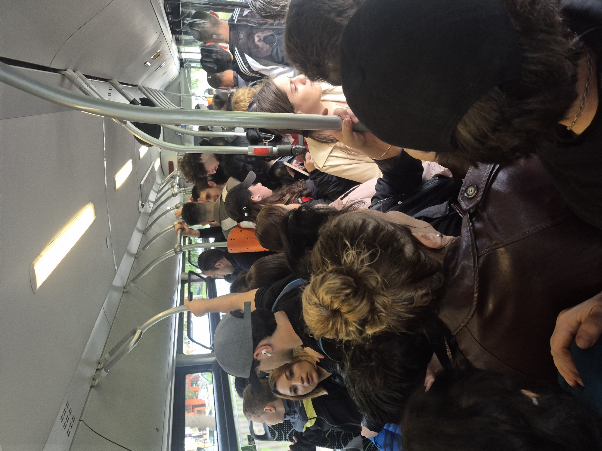 The interior of a crowded public bus in Tbilisi, Georgia is depicted. Passengers of varying ages and ethnicities are standing, closely packed together, and holding onto overhead handrails. The bus ceiling is white and has fluorescent lights. Outside the windows, buildings and trees are visible, indicating an urban environment.