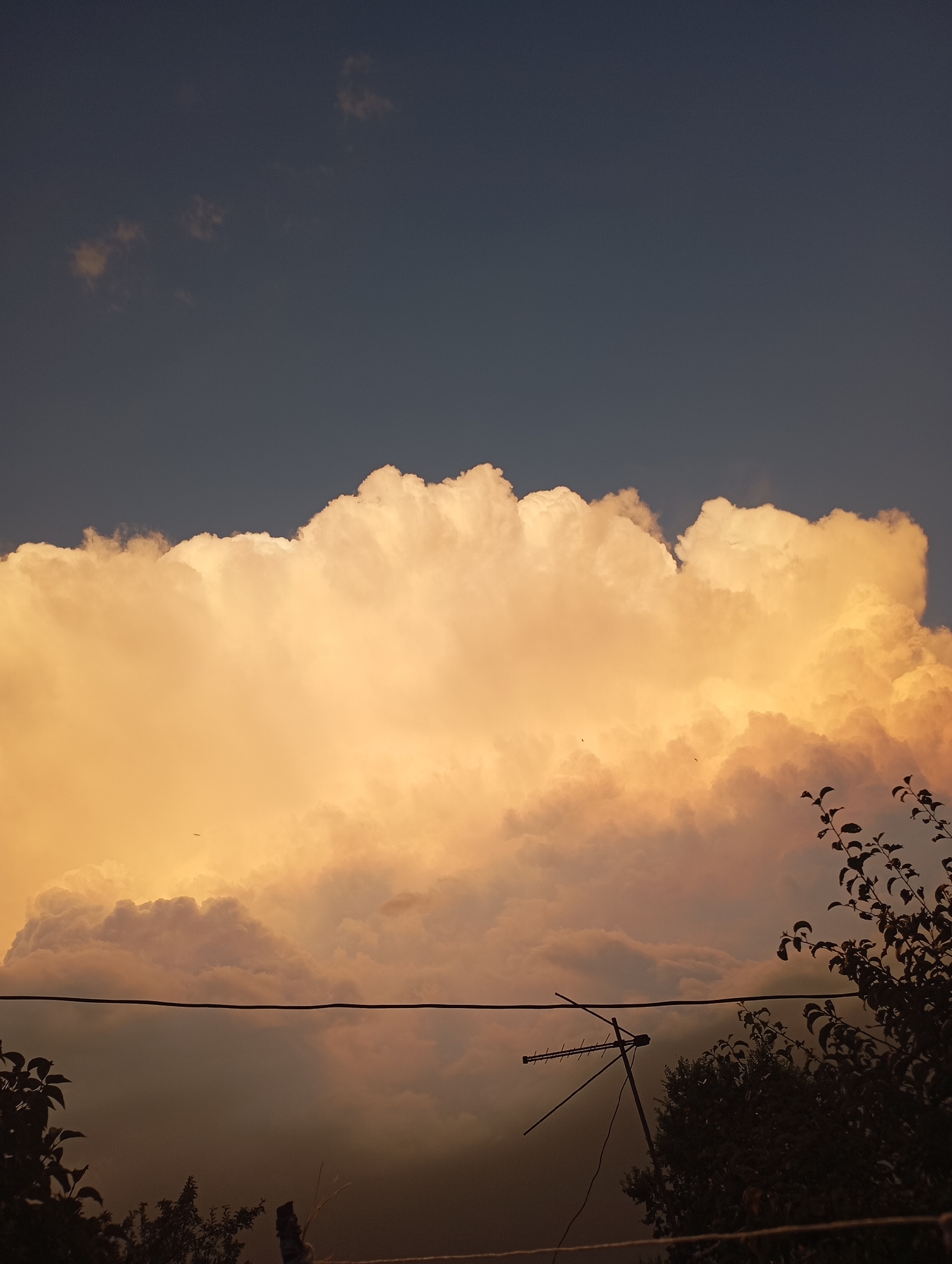 A dark sky is visible above a large, illuminated cloud formation. The clouds are a warm, golden color, suggesting they are lit by the sun. The bottom portion of the image is darker, with silhouettes of trees and a television antenna. A thin wire runs horizontally across the middle of the frame. The scene appears to be an outdoor shot, possibly taken at dusk or dawn, in the vicinity of Border Gyumri-Akhuryan, Armenia.
