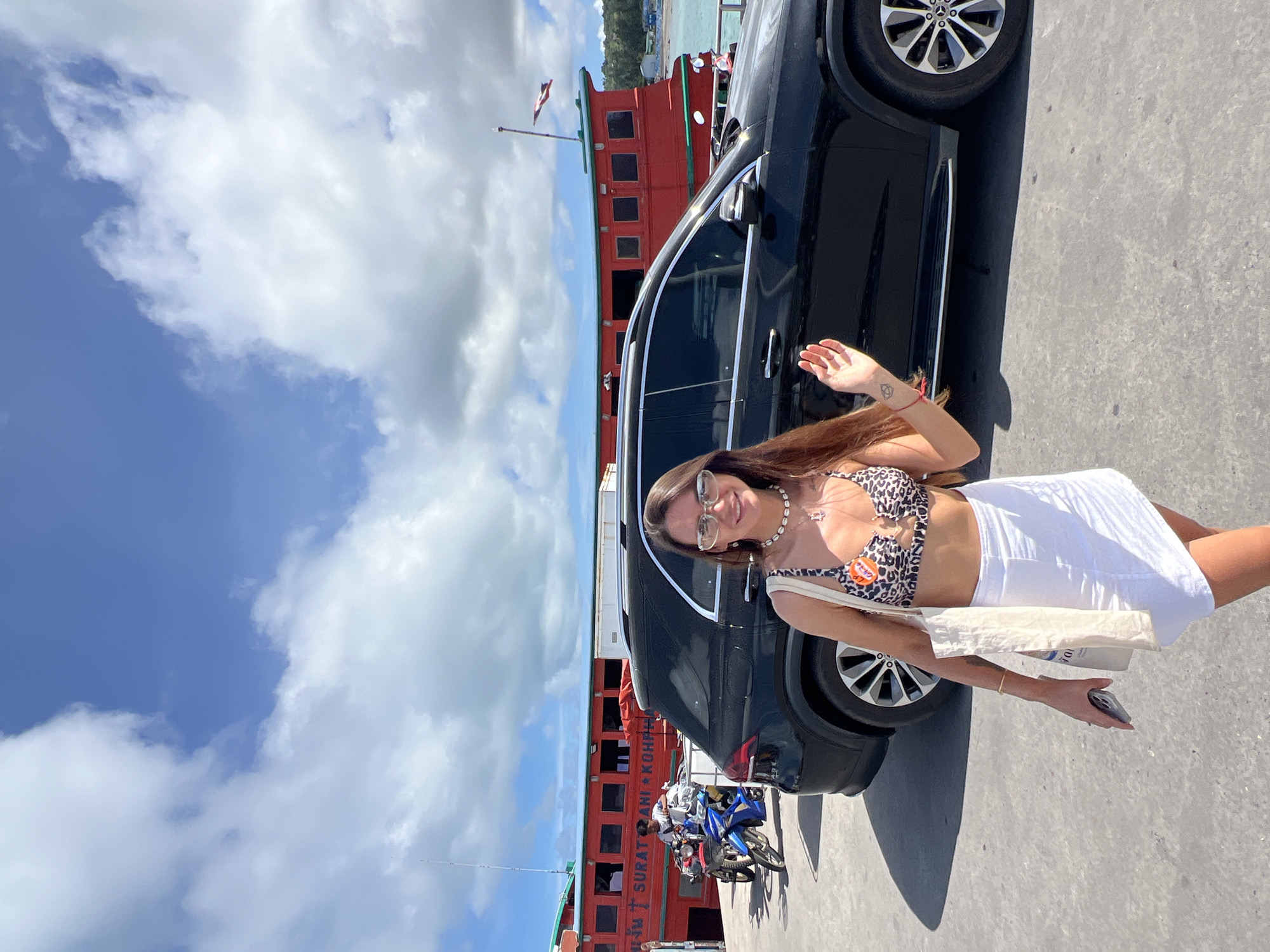 A woman stands on a concrete pier, smiling and waving at the camera. She is wearing a leopard print bikini top, a white skirt, and sunglasses. She holds a white tote bag in her left hand and a cell phone in her right. Behind her, a black SUV is parked, and further back, a large red ferry is docked. The ferry has "SURAT THANI KOH PHAN" written on its side. Two motorcycles are parked on the pier near the ferry. The sky above is blue with scattered white clouds. The scene appears to be at a ferry terminal on Koh Phangan island in Thailand.