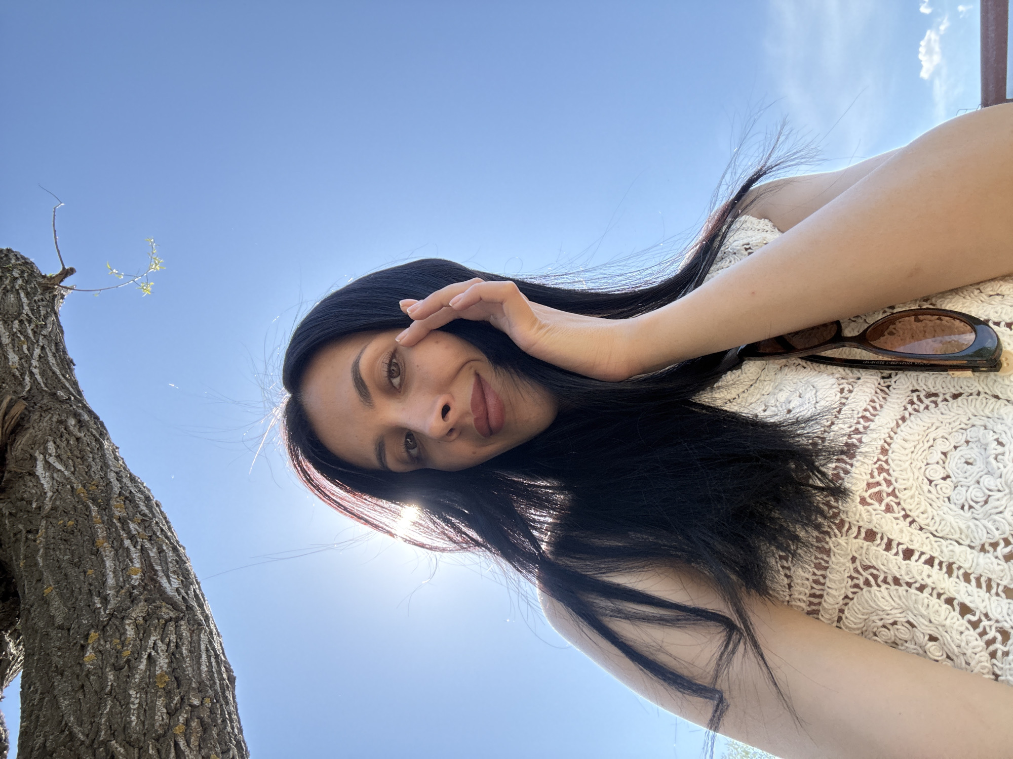 A woman is looking up at the camera with her hand near her face. She has dark hair, light skin, and is wearing a white patterned top. Next to her are sunglasses. Behind the woman is a blue sky, and part of a tree trunk is visible on the left side of the frame.
It is impossible to determine the exact location in Votkinsk, Russia from the image.