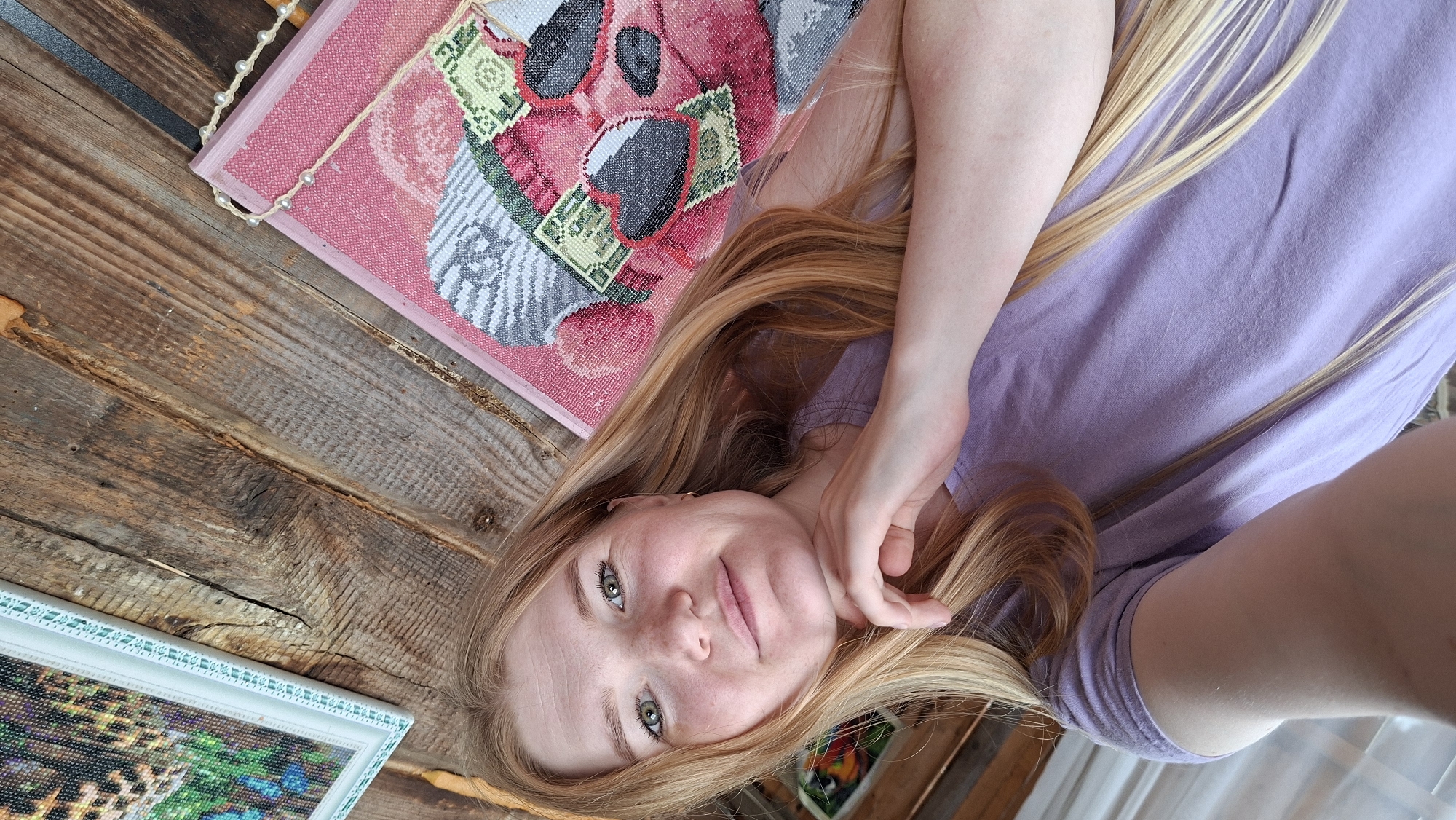 A young woman with long, blonde hair and green eyes lies on a wooden surface, looking towards the camera. She is wearing a light purple t-shirt and has her hand resting on her chin, with another hand near her hair. Her expression appears relaxed and happy. To her left, there is a piece of pink fabric with a colorful embroidered design featuring what looks like a bear or a panda wearing sunglasses and holding money. Below this, and to her right, is a framed picture with a colorful mosaic design, possibly a diamond painting. The wooden surface suggests an indoor or outdoor setting, possibly a rustic cabin or porch. The lighting appears to be natural, indicating it might be daytime. The overall mood is casual and peaceful. No visible text is present in the image.