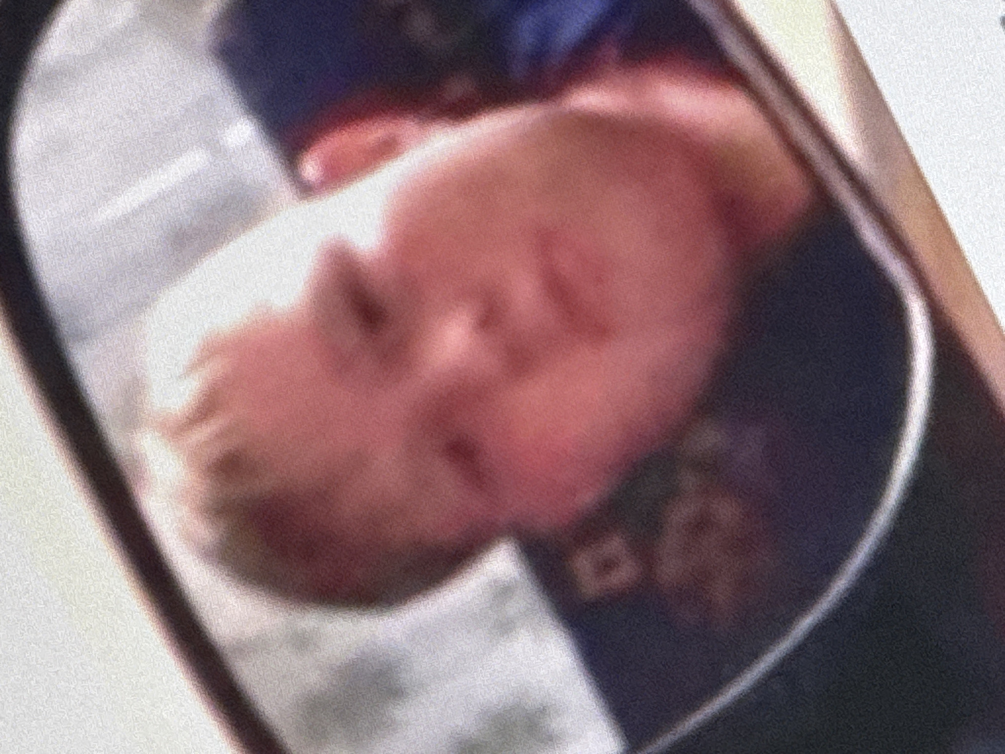 A close-up, slightly out-of-focus reflection in a mirror shows the face of a young boy, who appears to be Caucasian. He has fair skin, light-colored hair, and a round face with prominent cheeks. His eyes are visible, and his mouth is slightly open. The background of the reflection is dark, with some blurred objects that may be seats or upholstery. The boy's head is tilted slightly to the left. The lighting suggests an indoor setting, possibly a vehicle, as indicated by the curved frame of the mirror and the presence of what might be seating in the background. The image does not provide any specific information about the location in Saratov, Russia, or the context of the reflection.