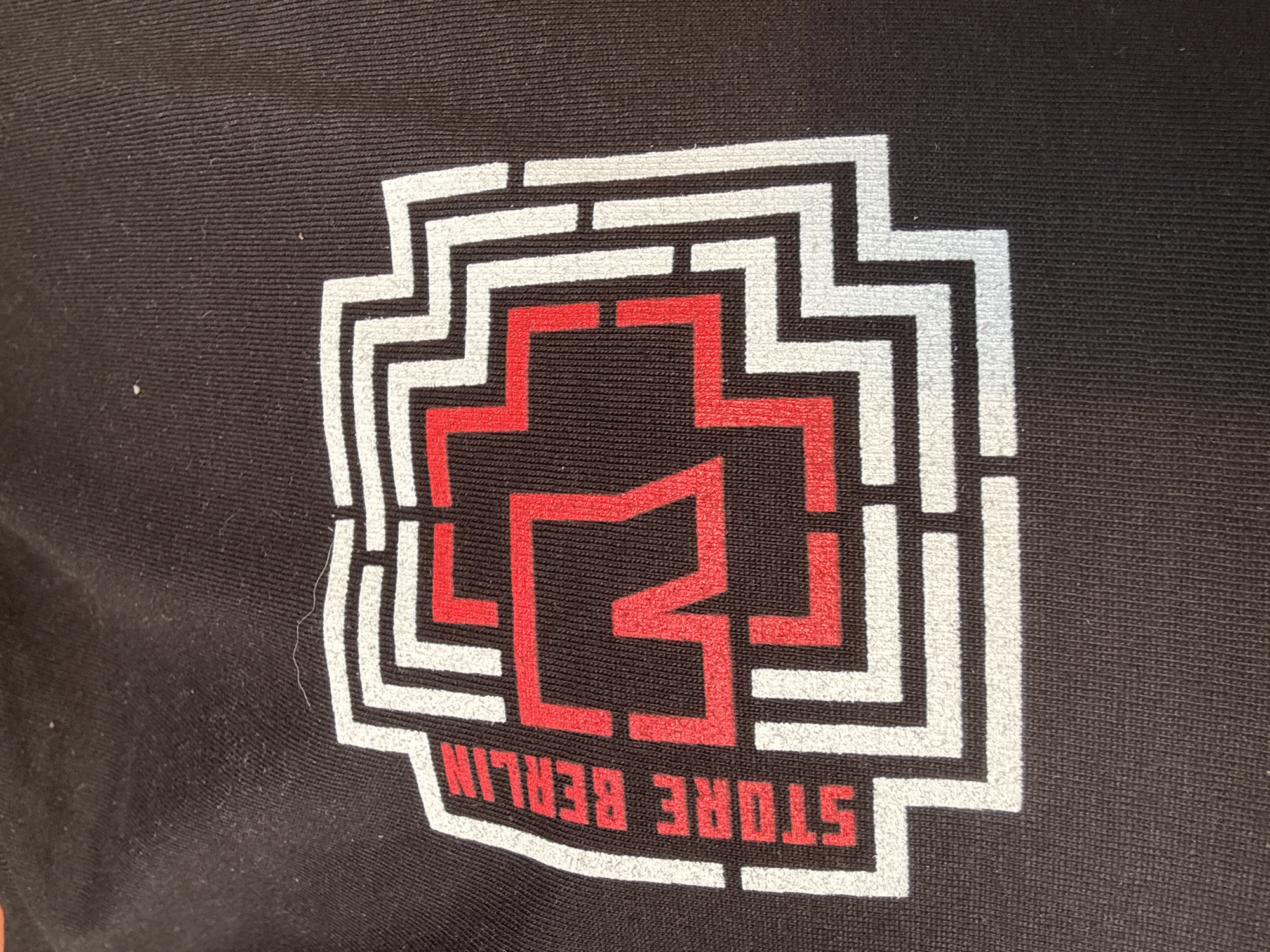 The image shows a close-up of a black fabric, likely a t-shirt, with a stylized logo printed in white and red. The logo is a geometric design with a central red cross-like shape surrounded by white, maze-like lines. On the left side of the logo, the white text "STORE BERLIN" is vertically oriented. The fabric exhibits a ribbed texture. No specific location in the city of Szczecin, Poland is discernible from the image.