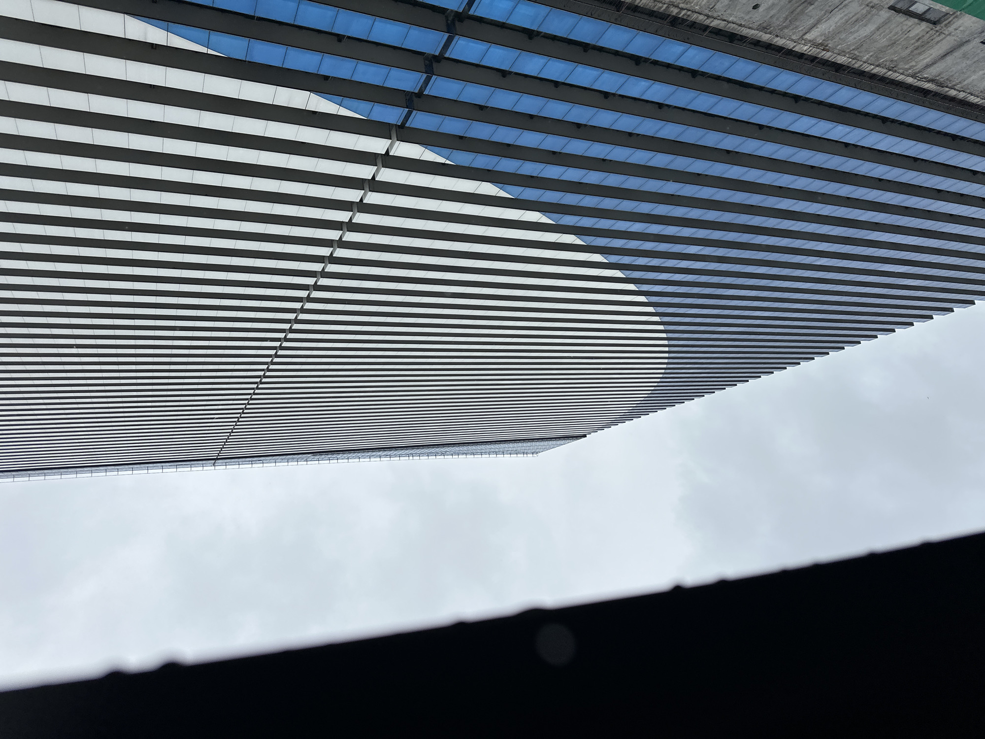 The image, taken from a low angle, depicts a building facade with horizontal white and dark stripes against a cloudy sky. In the upper portion, a section with a blue, grid-like structure is visible above the striped facade. A dark, out-of-focus object is in the foreground, likely a window frame or interior element. The location is potentially in Batumi, Georgia, given the presence of distinctive architectural elements commonly found in that city. Further details, such as identifying landmarks or building features, would require more specific information or closer proximity to known structures in the area.