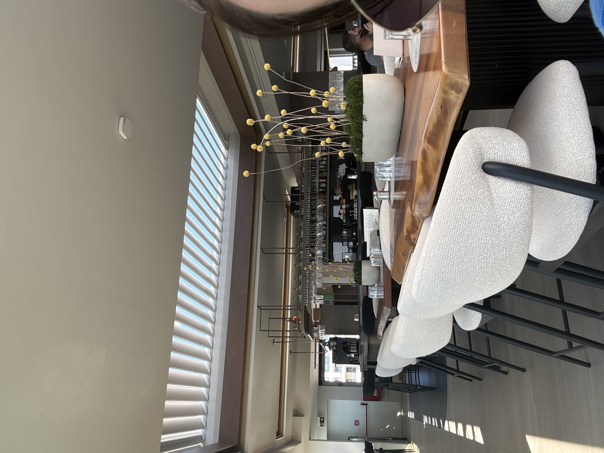 The image depicts the interior of a modern restaurant with a focus on a bar area and seating. A prominent feature is a long, polished wooden bar table with several upholstered chairs in front of it. A potted plant with tall, thin stems and yellow spherical flowers sits on the table. Sunlight streams through a slatted ceiling, casting striped shadows on the floor and chairs. In the background, a well-stocked bar with glassware and bottles is visible, along with some patrons. The overall atmosphere suggests a contemporary dining or drinking establishment. The specific location in Thermi, Greece, is not determinable from the visual information in the image.