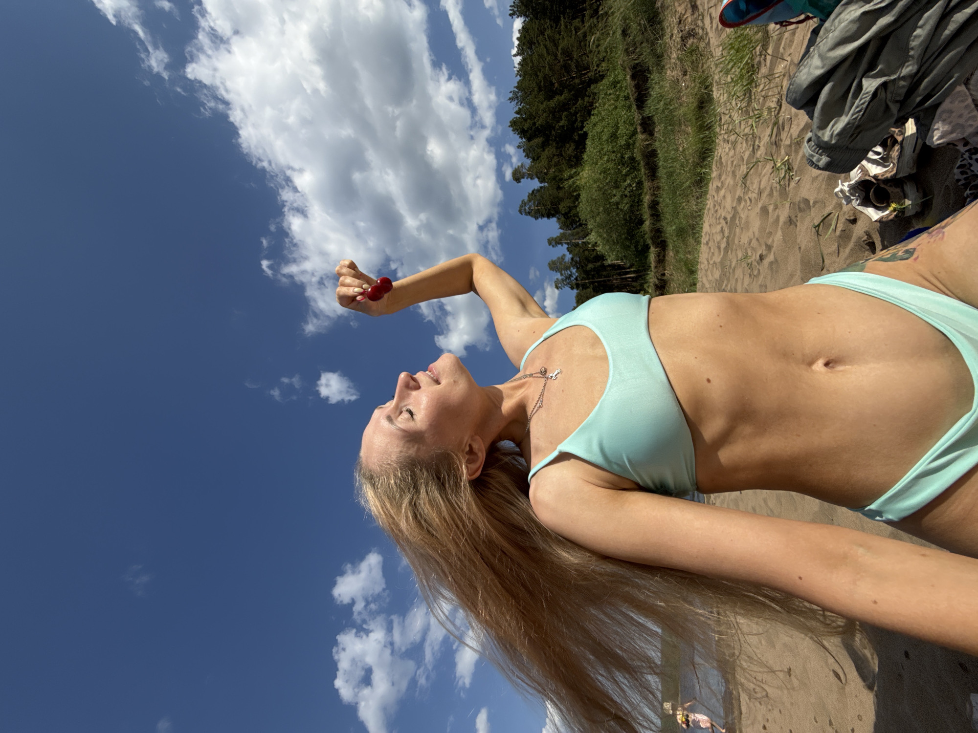 A blonde woman lies on a sandy beach against a backdrop of blue sky and clouds. She is wearing a light green bikini and holding a cherry in her outstretched hand. The location appears to be a beach near Chaikovsky, Russia, characterized by a mix of sand, vegetation, and trees along the shoreline. A pile of clothing and shoes lies in the upper right corner of the image.