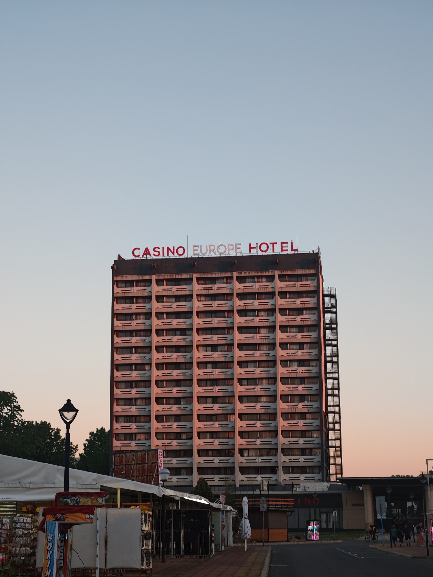 The image depicts the Casino Europe Hotel in Nesebar, Bulgaria, a multi-story building with a red sign on top. The building has a repetitive pattern of balconies and windows. A fire escape is visible on the right side of the structure. In the foreground, there are street-side kiosks or stalls, some appearing to be closed or covered. A lamppost stands to the left. A road stretches across the bottom of the frame, with a few people walking and cycling in the background. The sky is a pale blue color, indicating either early morning or late evening. The lighting casts a pinkish hue on the building's facade, suggesting the sun is setting or rising.