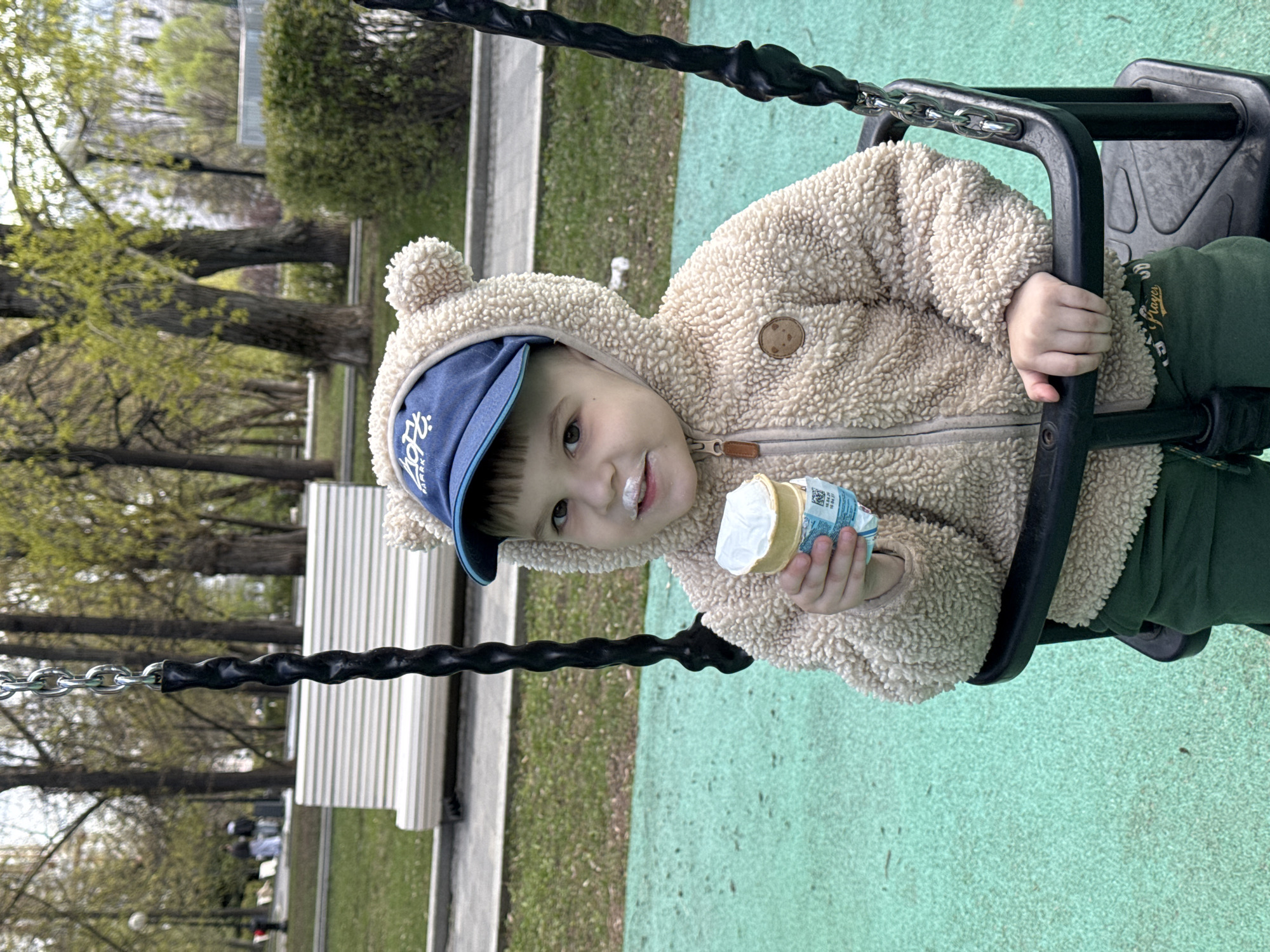 A young child with ice cream on their face sits in a black swing. The child wears a blue baseball cap, a cream-colored fleece jacket with a hood and bear ears, and green pants. They are holding an ice cream cone in their left hand. The background shows trees without leaves, indicating early spring. A park bench is visible in the background. Based on the style of the playground flooring and general environment, the location is likely a public park in Moscow, Russia.