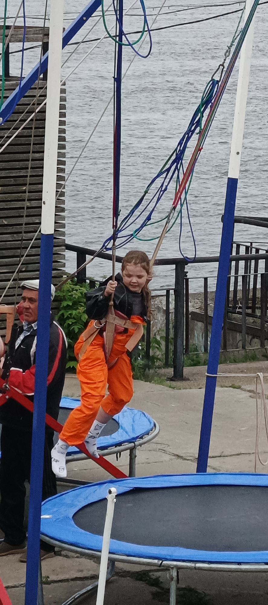 The image depicts a bungee trampoline activity in Sarapul, Russia, likely near the Kama River, given the body of water in the background. A young girl with light brown hair, approximately 5-8 years old, is the central figure, smiling while suspended in a black safety harness over an orange full-body suit and white patterned socks. Her arms are bent, and she is holding parts of the harness. Multiple colorful elastic cords (blue, green, red, black) attach to her harness and extend upwards, indicating she is in mid-bounce or elevated. To her left, an adult male, likely an operator or guardian, is partially visible, wearing a white flat cap, a black and white jacket, and dark pants, observing the girl.
The bungee trampoline system includes two prominent vertical poles: one white with a blue base on the left and a solid blue one on the right. A horizontal red bar is positioned below the girl's feet. Two trampolines with blue protective padding and dark jumping surfaces are on the concrete ground, one partially beneath the girl and another to the right.
In the background, a body of water with ripples is visible. To the far left, a wooden slatted structure extends towards the water. To the right of the girl, a concrete or stone embankment with black metal railings separates the activity area from the water. Green foliage is present behind the man and the girl. The overall lighting suggests an overcast sky.