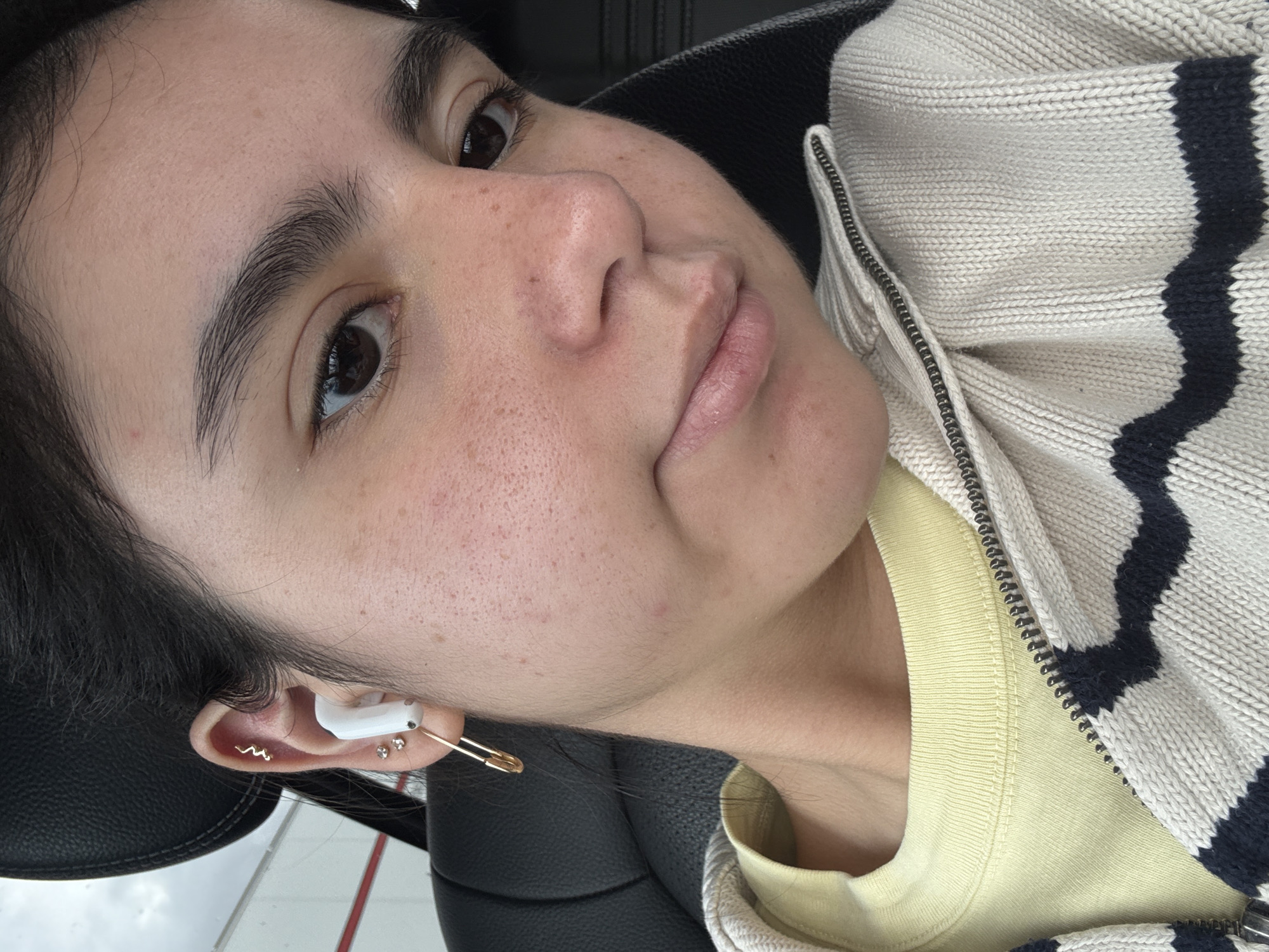 A close-up selfie depicts a light-skinned person with dark hair wearing a light yellow t-shirt and a beige and navy striped knit zipper jacket. The person has brown eyes, full lips, and visible freckles. They are wearing a white wireless earbud and a gold safety pin in their left ear. The person appears to be seated in a black leather seat. The background is slightly blurred, showing what appears to be a building exterior. The location, San Marcos, United States of America, is inferred from the prompt.