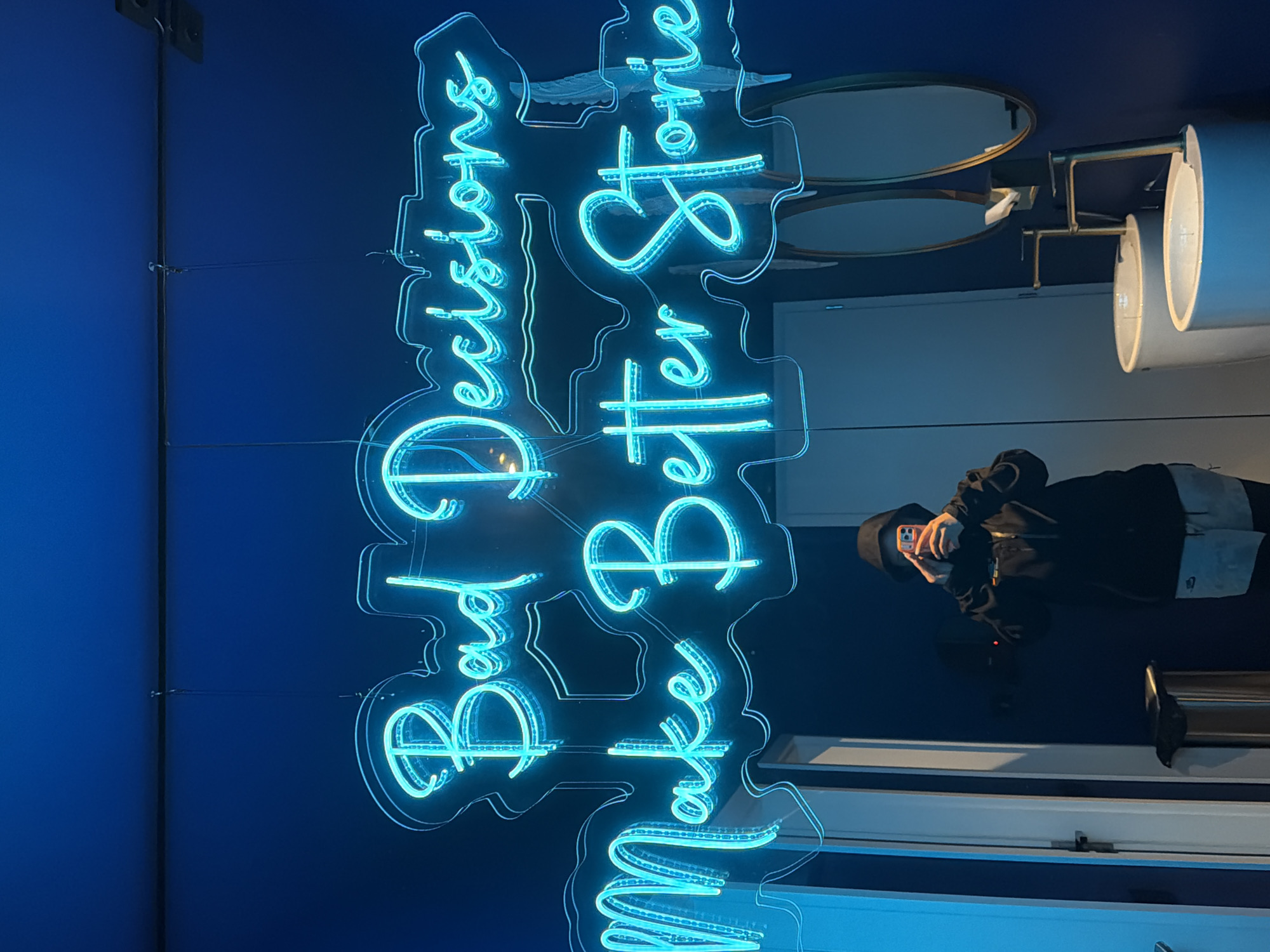 The image depicts an indoor scene, oriented 90 degrees counter-clockwise from its upright position. The central focus is a vibrant blue neon sign, glowing against a deep blue wall. The sign displays the cursive text: "Make Better Love & Bad Decisions".
Below the neon sign, a person is reflected in a glossy, dark surface, seemingly taking a selfie with their phone. The person is wearing a dark hooded jacket and a dark hat or cap, holding a phone with a light-colored case.
To the right of the neon sign, several decorative elements are visible. These include two oval-shaped mirrors with slender gold or brass frames, stacked vertically. Below the mirrors, two cylindrical light fixtures, possibly wall sconces, also featuring gold or brass accents, are mounted on the wall.
The lighting in the room appears dim, emphasizing the bright glow of the neon sign. The overall color palette is dominated by various shades of blue, complemented by the metallic gold/brass tones of the decor and the dark attire of the reflected person. The setting, given the neon sign and stylish decor, suggests a modern interior space like a bar, cafe, or lounge. The location context provided is Sofia, Bulgaria.