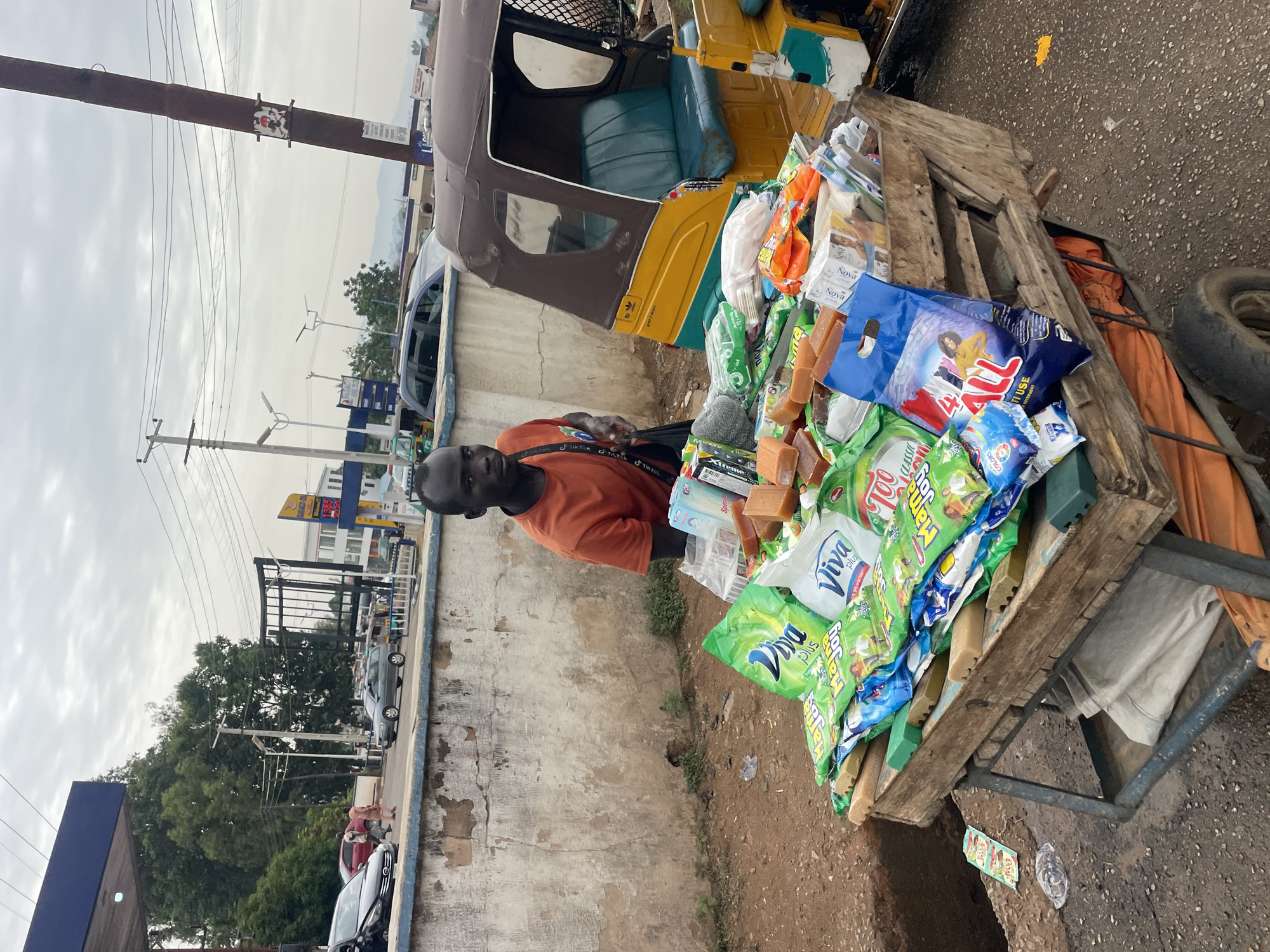 The image depicts a street vendor in Jos, Nigeria. The vendor, a Black man wearing an orange shirt, stands next to his makeshift cart laden with goods. The cart is constructed from a wooden pallet and a metal frame, filled with various cleaning products, soap bars, and other household items. A yellow and brown auto-rickshaw, also known as a keke, is parked adjacent to the cart. The vendor has a bag slung across his shoulder with a repeated "TikTok" logo on the strap. The background shows a concrete wall, a street scene with cars, power lines, and buildings. The sky is overcast. The location appears to be a busy street corner.