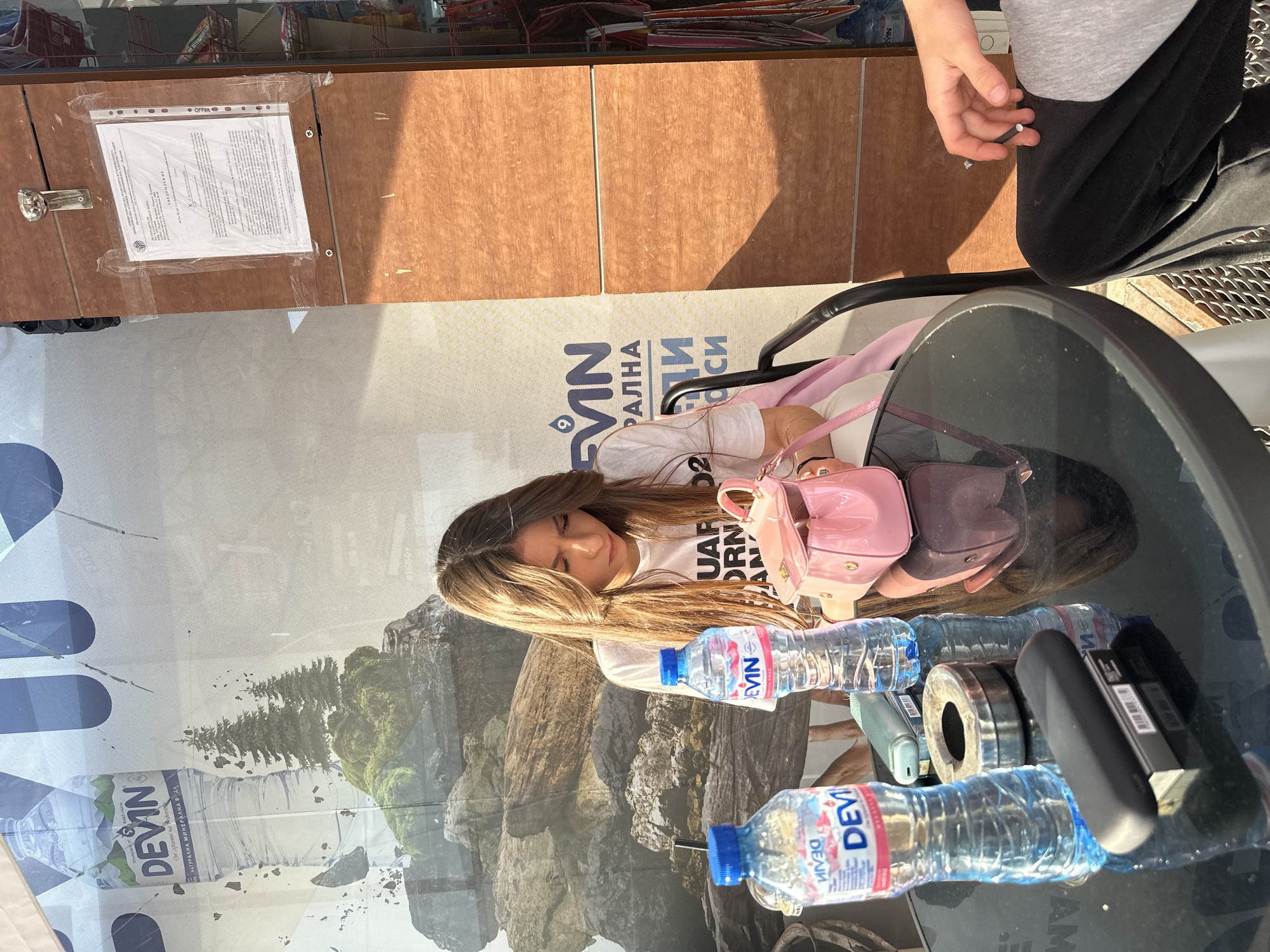 A young woman with long, light brown hair sits at a reflective, dark-surfaced table in what appears to be an outdoor or semi-outdoor cafe setting in Plovdiv, Bulgaria, during bright daylight. She is wearing a white t-shirt and has a pink handbag on her lap, with her gaze directed downwards. On the table are two plastic bottles of DEVIN brand mineral water, a black electronic device, and a metallic ring. Behind her, a large banner displays a natural mountain landscape with pine trees and prominent text including "DEVIN" and Bulgarian words like "НАТУРАЛНА МИНЕРАЛНА ВОДА" (Natural Mineral Water) and "От извора на Водоп" (From the source of Vodop). Partially visible in the upper right is another person's hand holding what resembles a cigarette or vape. Above the banner, on a wooden wall, a document with "OFFIM." printed at the top and extensive Cyrillic text is taped, possibly an official notice. The scene is calm and casual, illuminated by strong sunlight.