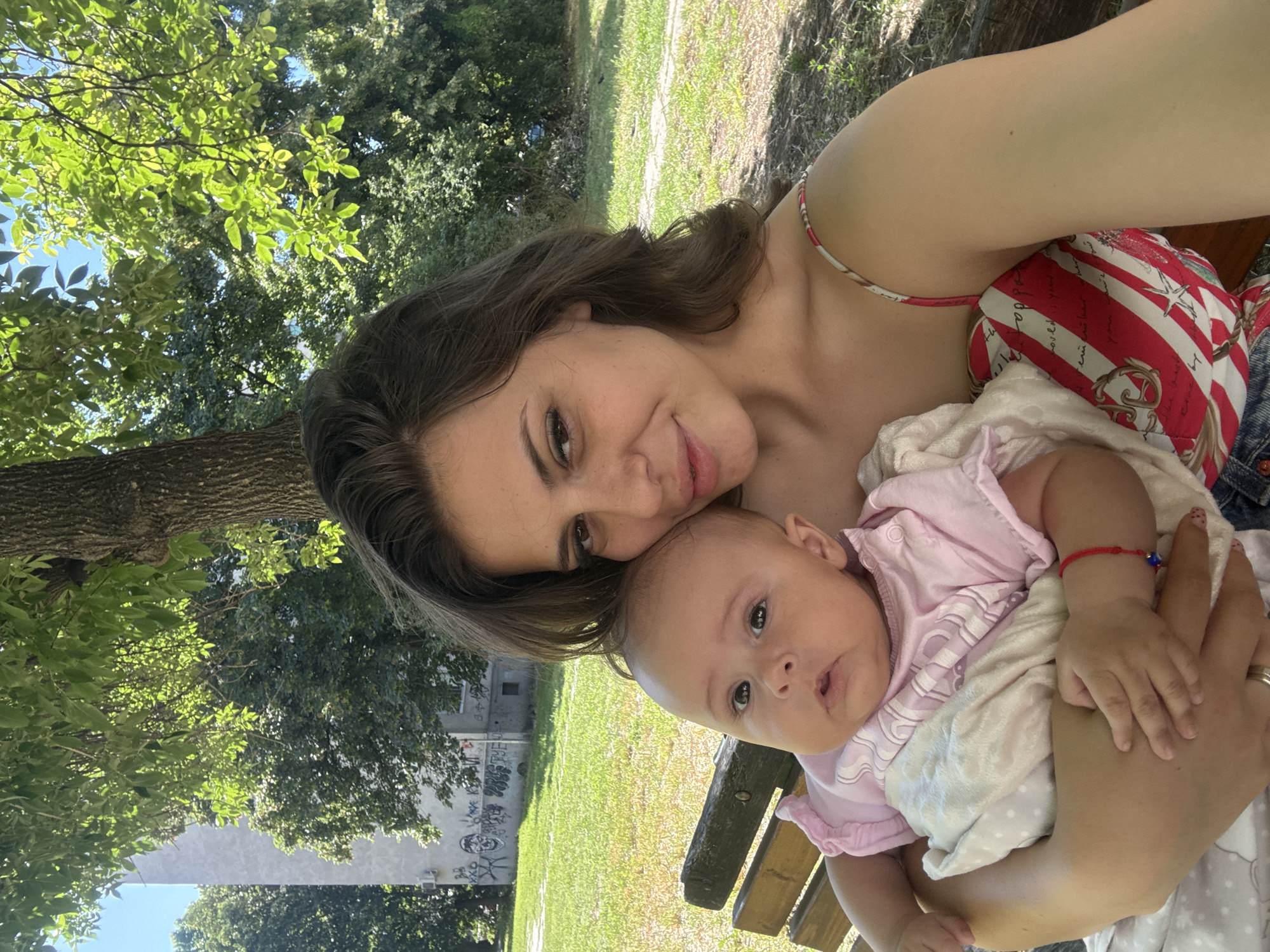 In Plovdiv, Bulgaria, a woman is holding a baby on a wooden park bench. The woman has brown hair and is wearing a red and white striped top. The baby is wearing a pink outfit. In the background, there is a building with graffiti on it, trees, and grass. The overall environment suggests a sunny day in a park.