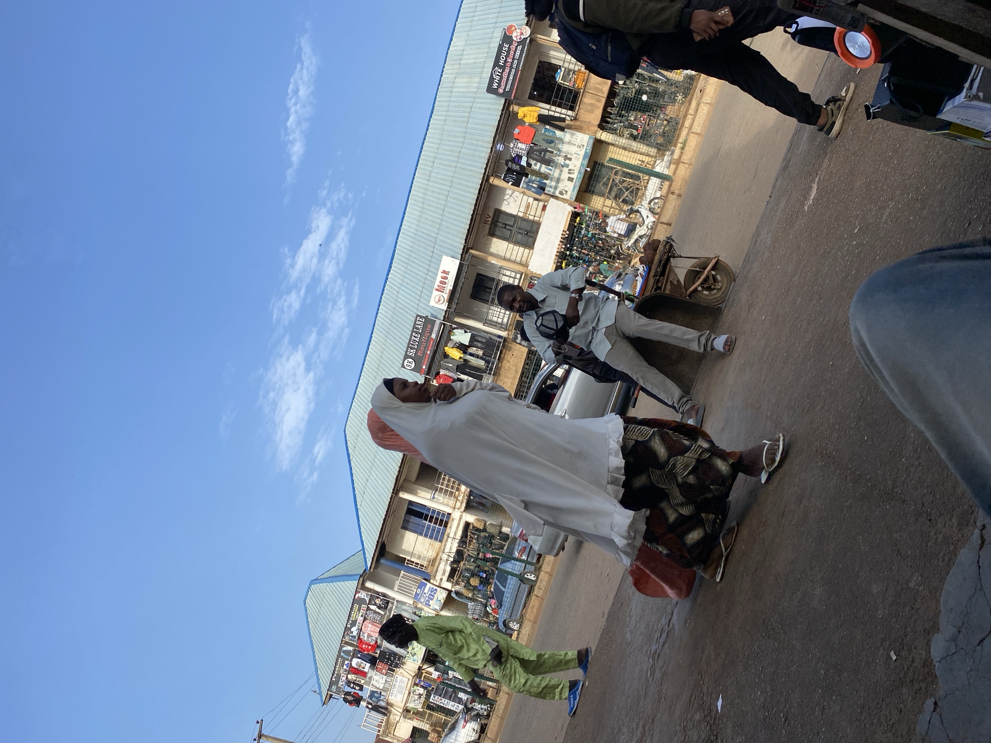 The image captures a street scene in Bukuru, Nigeria, during daylight hours. The sky is clear blue, with a few clouds. The scene features several people, including a woman wearing a white headscarf and a patterned dress, carrying a red bag. A man in a light-colored shirt and trousers is standing near a cart filled with produce. Another person is wearing a green outfit and walking in the opposite direction. On the right side of the frame, an individual is partially visible, wearing dark clothing and shoes.
Several storefronts are visible in the background, some with signage such as "SK LUXE LANE Boutique," "Rv 90 00," "WHITE HOUSE Cosmetics & Jewelry," and "Mock." Merchandise is displayed outside some of the stores. Vehicles are parked along the street. The overall environment is a typical urban street in Nigeria.
