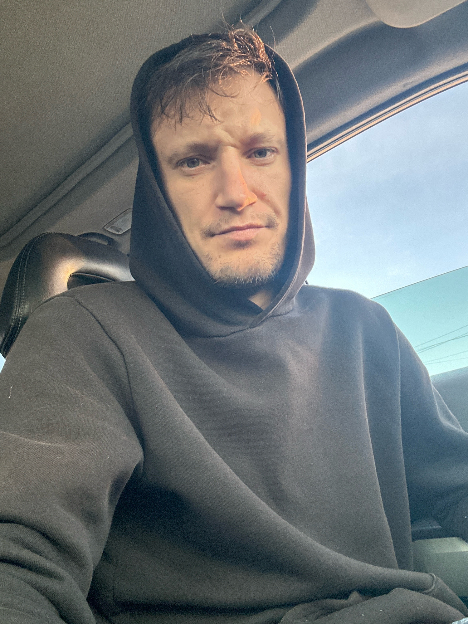 A selfie taken inside a vehicle shows a man with light brown hair and stubble wearing a dark hooded sweatshirt. The man is looking directly at the camera with a slight grimace. The interior of the vehicle is visible, including a dark headrest and the car's ceiling. Sunlight is visible in the background, suggesting it is daytime. The image does not provide any specific details that would indicate the location in the city of Makiv, Ukraine.