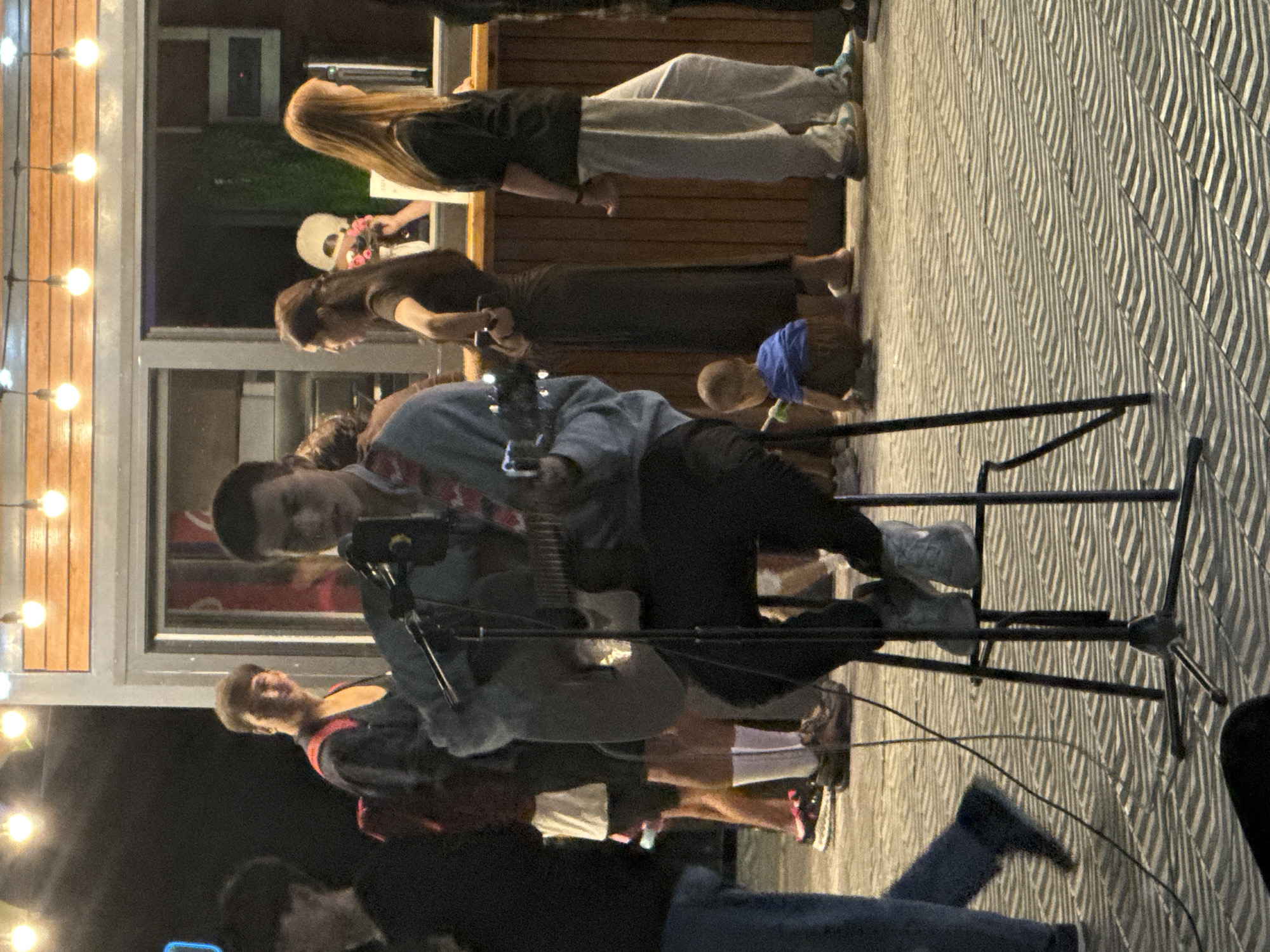 In Pavlodar, Kazakhstan, on a paved outdoor area with a patterned surface, a group of people are gathered. In the foreground, a young man sits on a stool, playing an acoustic guitar and singing into a microphone. He is wearing a grey sweatshirt and dark pants. Behind him, other individuals are standing and conversing, some near a food stall or kiosk with bright overhead lights. To the left, a person in a dark shirt and blue jeans is visible. Further to the right, a child in a blue shirt is seated on the ground. The scene appears to be a casual outdoor gathering or performance.