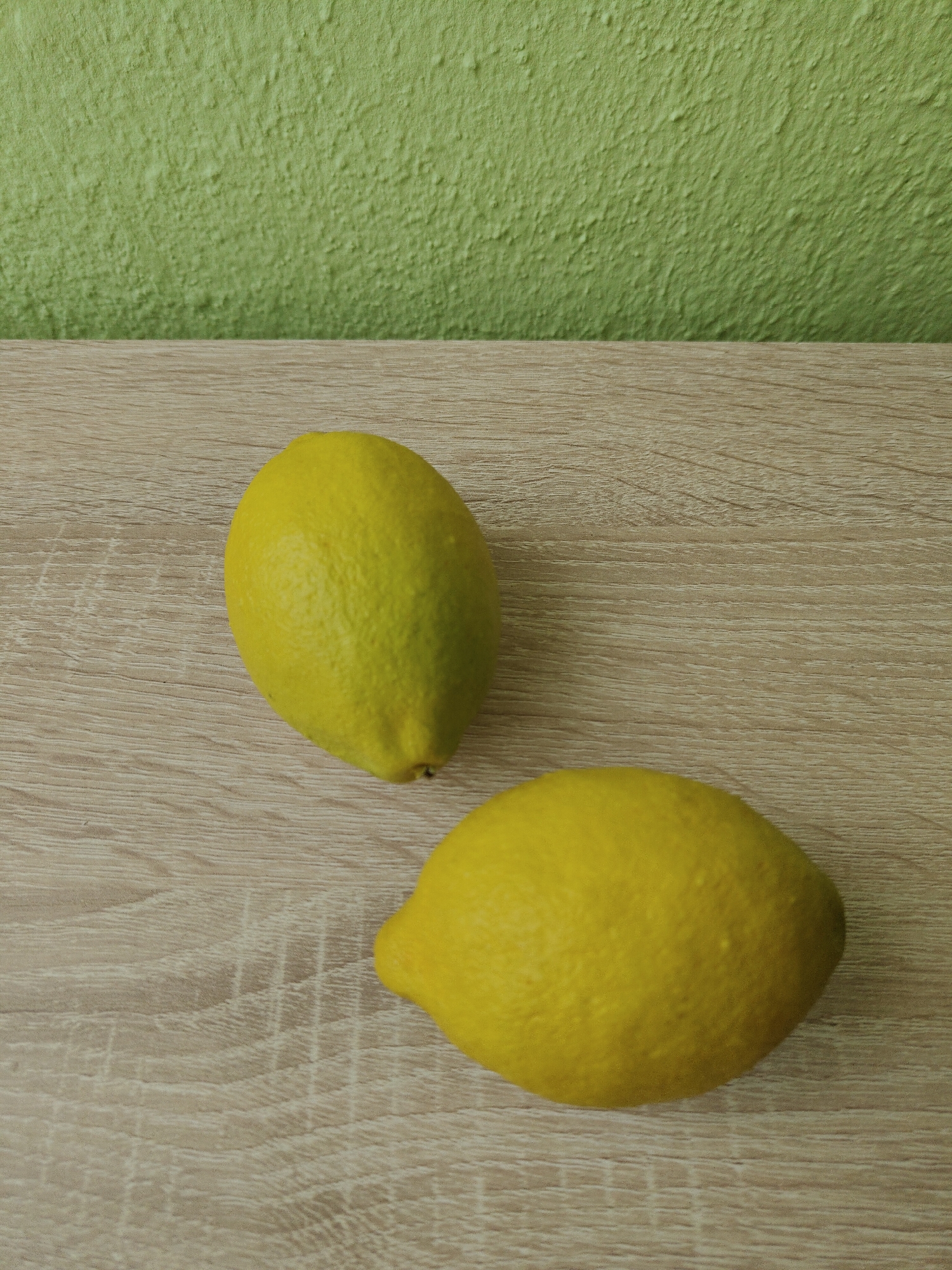 The image shows two lemons placed on a light brown, wood-grain-patterned surface. The background consists of a light green wall with a textured surface. The scene appears to be indoors. There is no information about the city of Unknown, Unknown.
