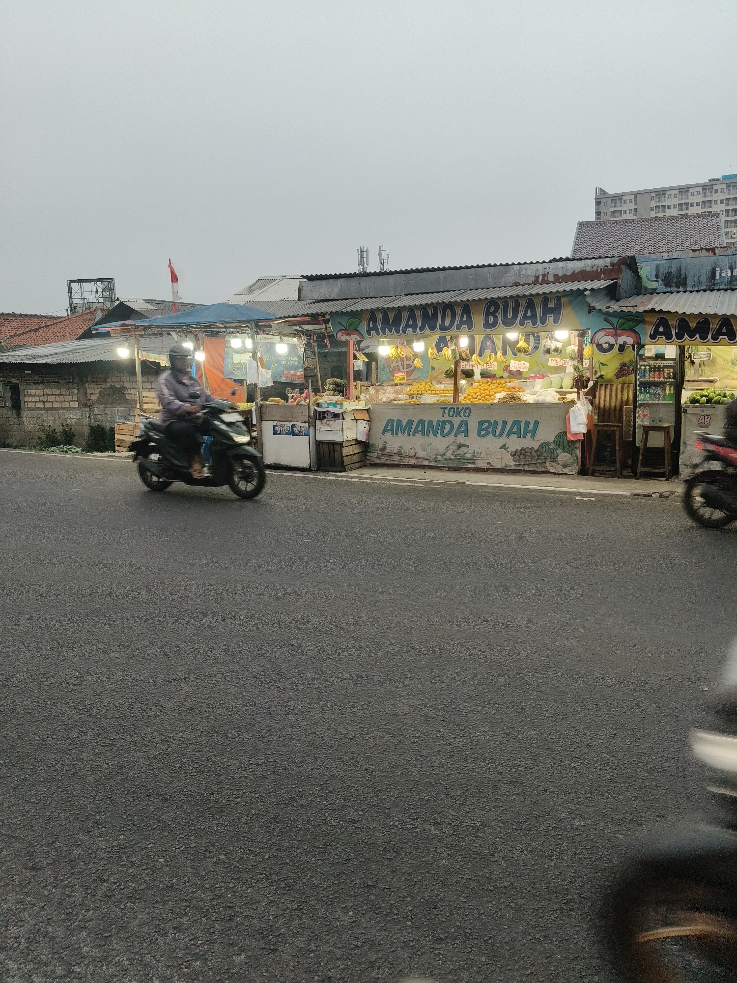 The image shows a street-side fruit stall in South Tangerang, Indonesia, as evidenced by the sign "TOKO AMANDA BUAH" (Amanda Fruit Shop) and the Indonesian flag visible on a pole above the stall. The stall is illuminated by bright lights, showcasing an assortment of fruits such as oranges, bananas, melons, and grapes. Bottles of drinks are also displayed. In the foreground, a person is riding a dark-colored motorcycle on an asphalt road, moving from left to right. Another motorcycle is partially visible on the far right side of the image, its rear wheel blurred by motion. The environment appears overcast, with buildings and a taller structure visible in the background, suggesting an urban or semi-urban setting. The overall scene depicts a typical street vendor operation in a bustling Indonesian city.