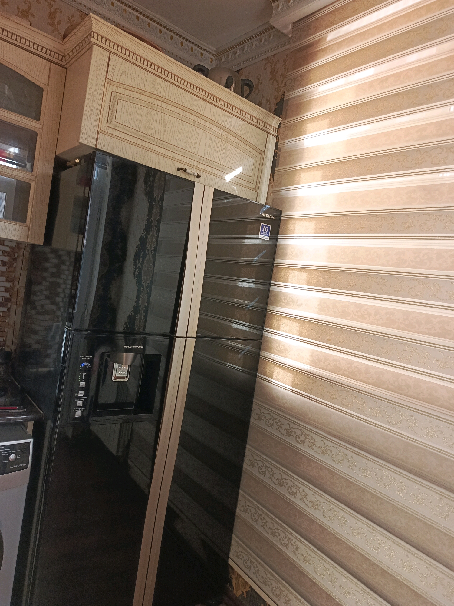 The image depicts a kitchen interior, likely in Khyrdalan, Azerbaijan. A large, black, two-door refrigerator with an inverter and a water dispenser is the central object. It stands in front of a wall covered in striped wallpaper with a subtle floral pattern. Above the refrigerator, light-colored kitchen cabinets with decorative molding are installed. To the left of the refrigerator, a portion of a washing machine is visible. The scene appears to be indoors, possibly in a residential setting, with artificial lighting casting shadows.