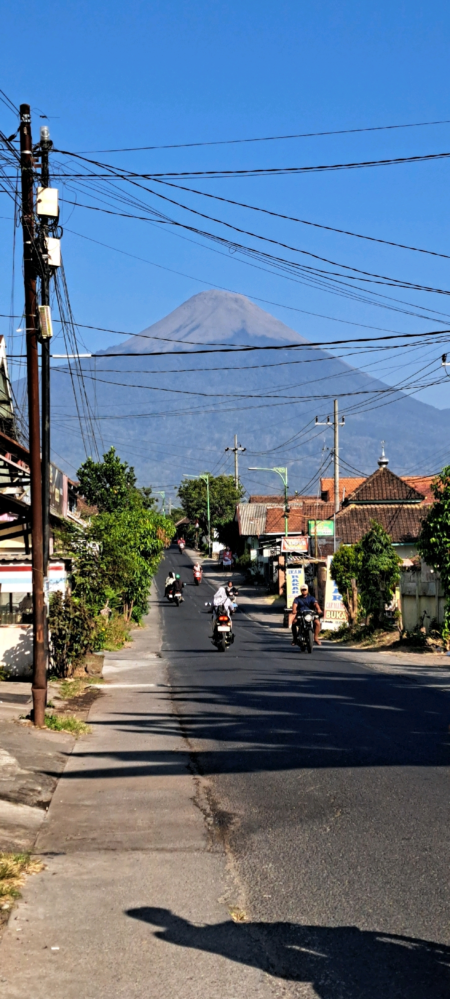 The image captures a street scene in Sumber Gedang, Indonesia, dominated by a vast, hazy blue mountain in the background under a clear, bright blue sky. The foreground shows a paved road with several motorbikes in motion, carrying individual riders. Some riders are wearing helmets, while others are not. The road is lined with buildings on either side, some with tiled roofs, and lush green trees. A prominent wooden utility pole with numerous black wires crisscrossing the frame stands on the left side of the road. Shadows of trees and possibly pedestrians are cast across the road and sidewalk. The overall impression is a typical daytime scene on a moderately busy street in a tropical or subtropical location.