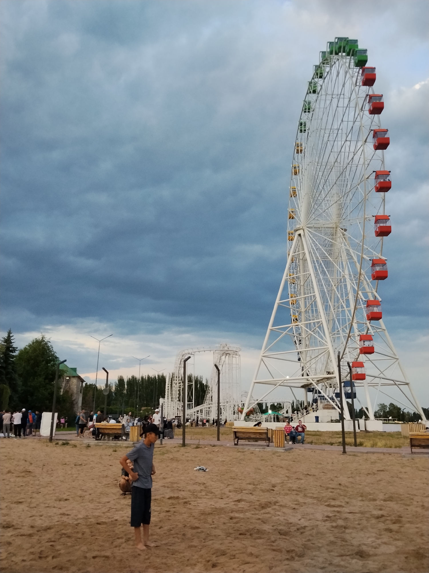 This media file depicts an amusement park scene on an overcast day. The most prominent object is a large, white Ferris wheel with red and green gondolas, positioned to the right of the frame. To the left of the Ferris wheel, a white roller coaster structure is visible.
In the foreground, a sandy area is dotted with people. A young boy wearing a gray t-shirt and dark shorts stands in the center, looking away from the camera. Further back, several groups of people are scattered throughout the park, some sitting on benches, others standing and conversing. Some individuals appear to be walking around.
The setting includes trees and buildings in the background, suggesting a recreational area. The sky is filled with dark, heavy clouds, indicating the possibility of rain or a storm. The overall mood seems relaxed, with people enjoying the park despite the weather. The time of day is difficult to determine precisely due to the cloud cover, but it appears to be late afternoon or early evening. No visible text is present in the image.