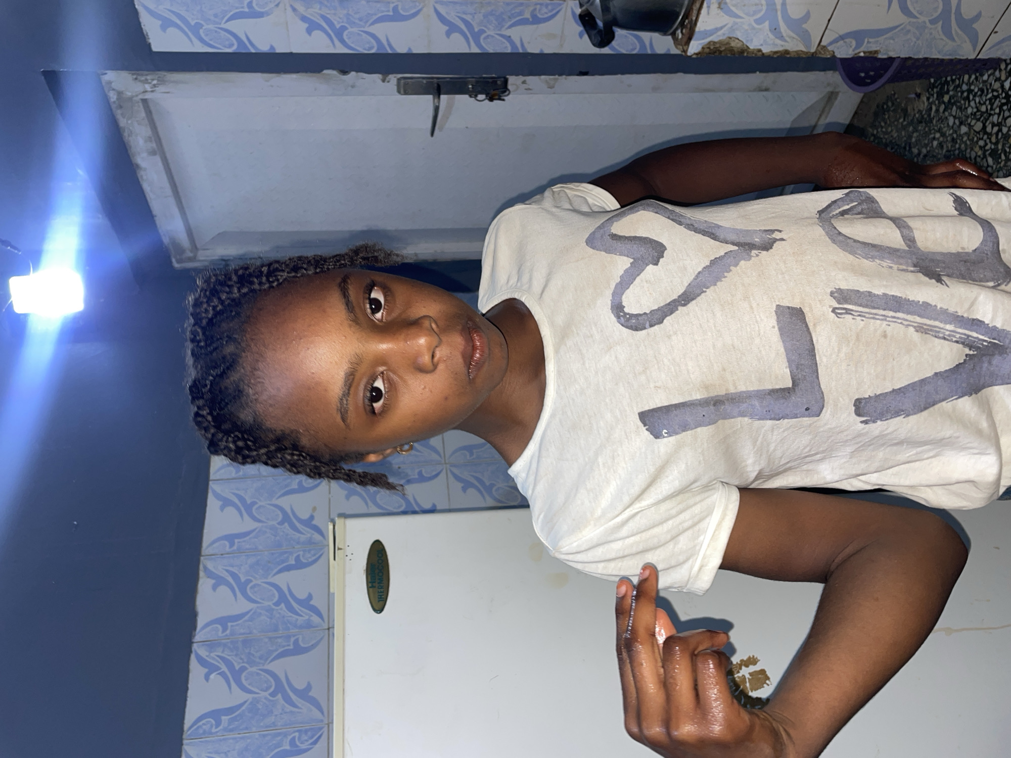 A young woman is centered in the frame, wearing a white t-shirt with "LOVE" printed on it. She is looking directly at the camera with a neutral expression. Her hair is styled in braids. Her right hand is raised, with what appears to be oil or a similar substance on it.
The background shows a kitchen setting with blue and white patterned tiles on the walls. A white refrigerator is visible behind her on the right.
The location is implied to be Jos, Nigeria.
