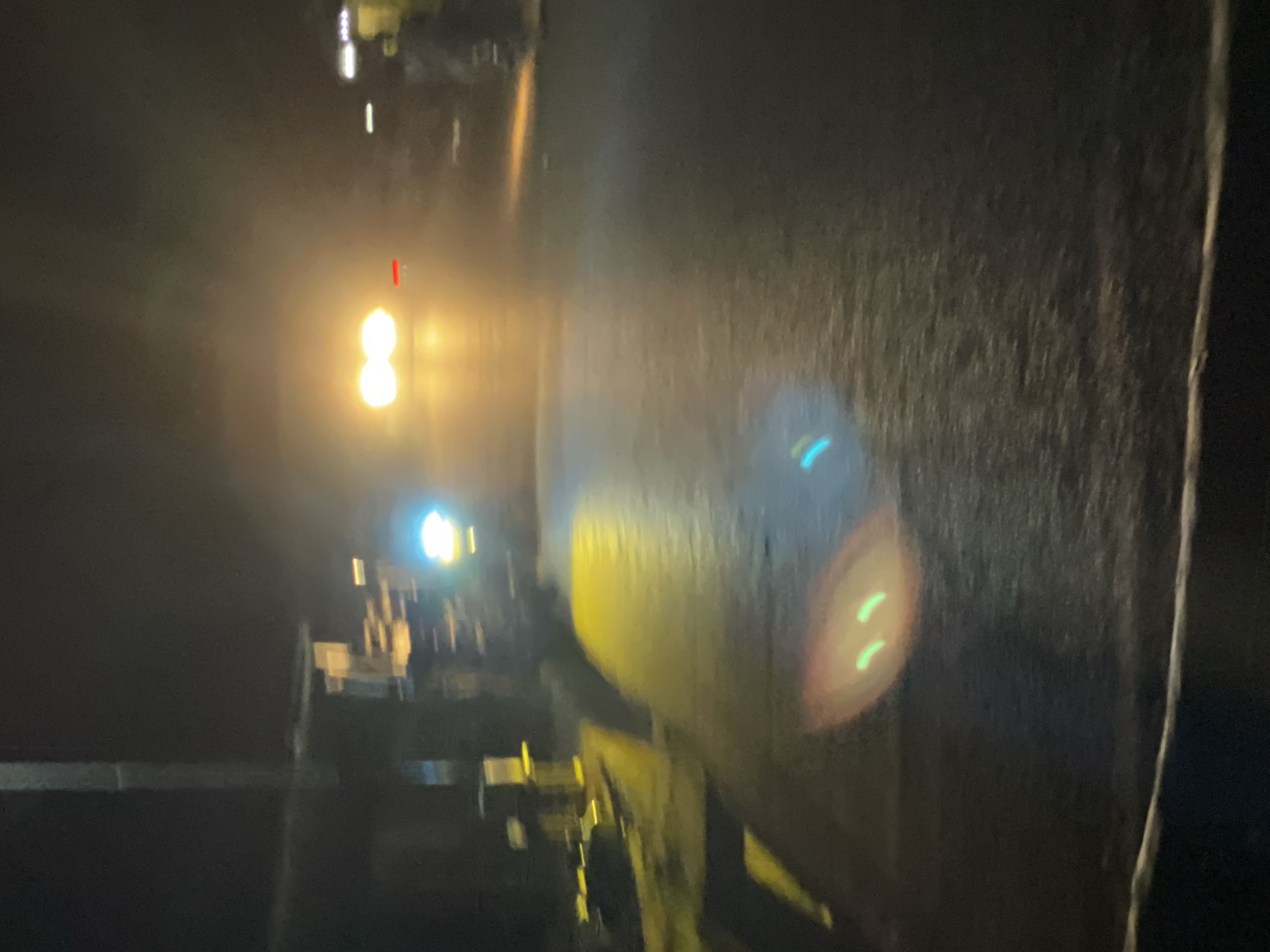 The image shows a dark, possibly nighttime, scene. In the distance, there are two bright lights, possibly headlights of a vehicle or streetlights. To the right, there's a dark, textured surface. The scene appears to be on a street or road, and the presence of lights suggests it's a developed area. The picture is blurry. Location cannot be determined solely from the image without additional clues.